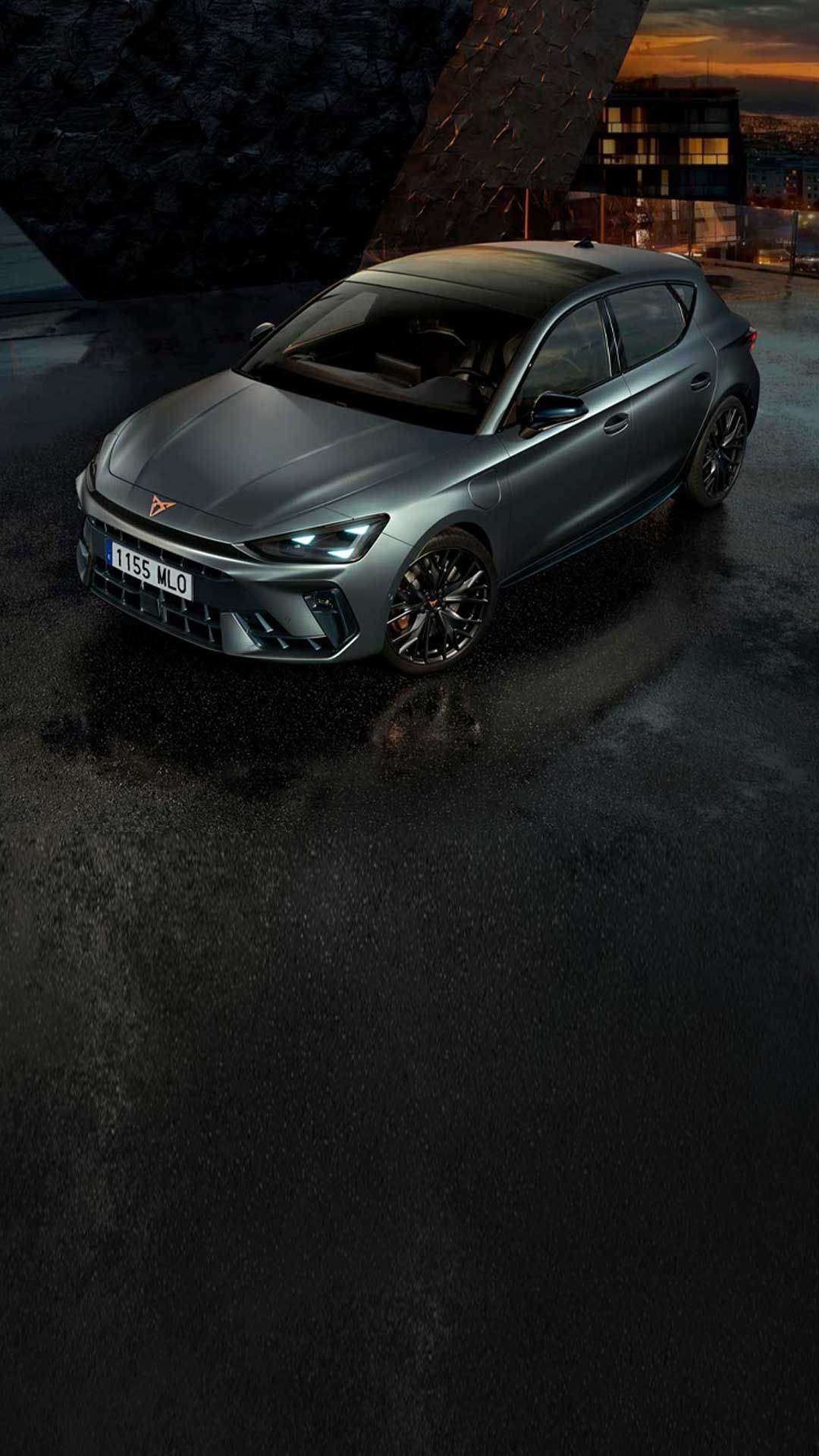 CUPRA Leon v kovinsko sivi barvi stoji na mokri strešni terasi ob mraku, v ozadju se blešči razsvetljeno mesto. 