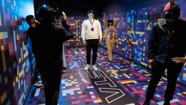 V skupini ljudi v prostoru z barvitimi pikselnimi vzorci na tleh in stenah preizkuša virtualna očala.