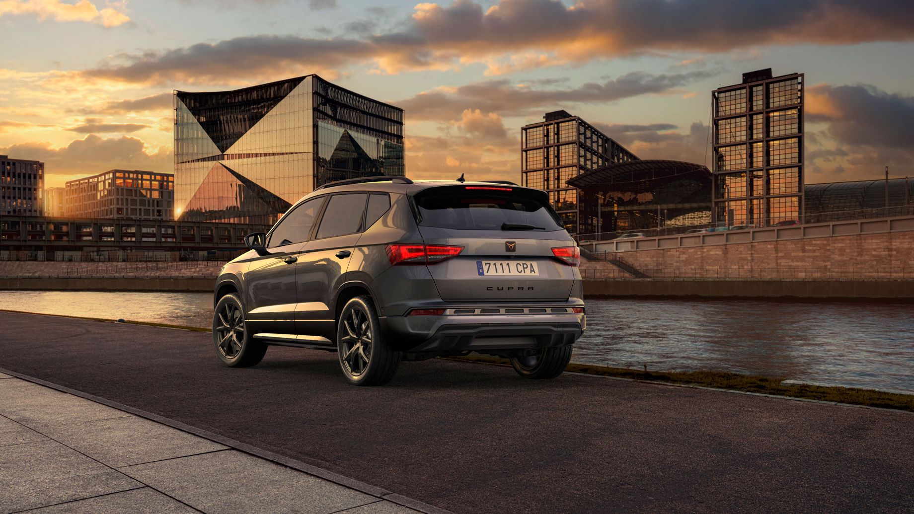 Parkiran CUPRA Ateca ob reki ob sončnem zahodu. V ozadju so sodobne steklene zgradbe in nebo z oblaki. Vozilo je obrnjeno stran od kamere proti reki.