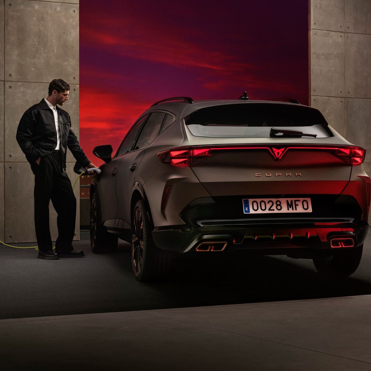 Moški polni električni CUPRA Formentor, ki je parkiran ob steni z električno polnilnico. V ozadju je videti dramatičen sončni zahod.