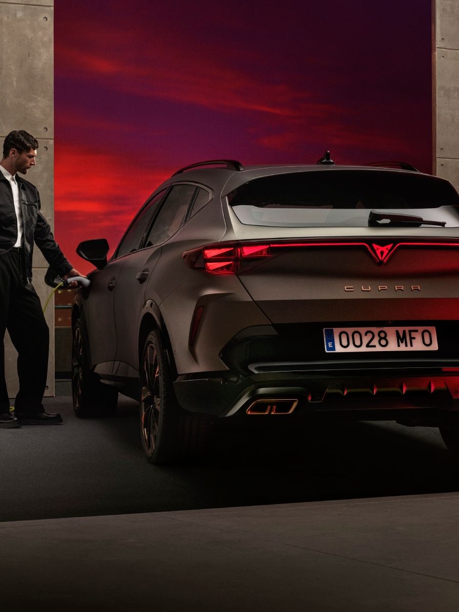 Moški polni električni CUPRA Formentor, ki je parkiran ob steni z električno polnilnico. V ozadju je videti dramatičen sončni zahod.