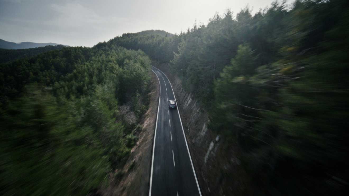 Pogled iz zraka na avtomobil, ki vozi po cesti skozi gost gozd. Okolica poudarja občutek svobode in povezanosti z naravo.