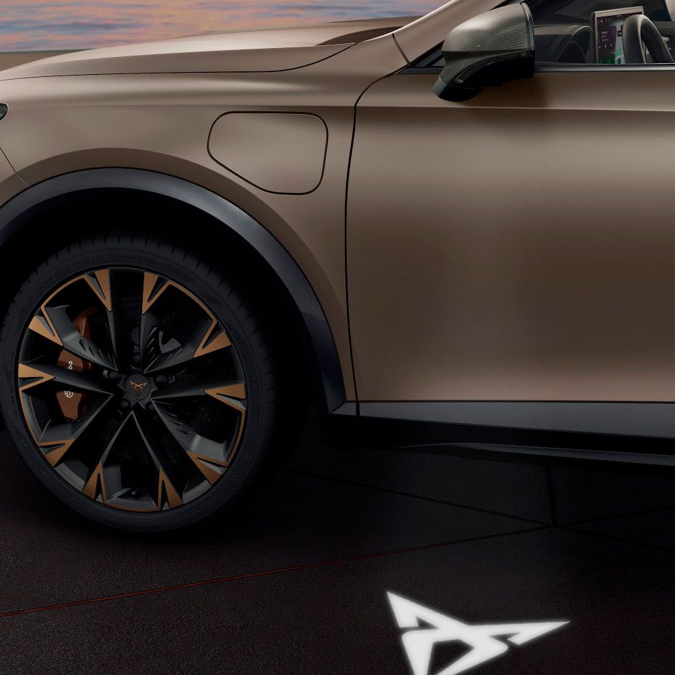 Sprednji del avtomobila CUPRA Formentor je prikazan s poudarkom na sprednjem kolesu. Vozilo je mat svetlo rjave barve, parkirano na pločniku s svetlečim emblemom na tleh pod avtomobilom.