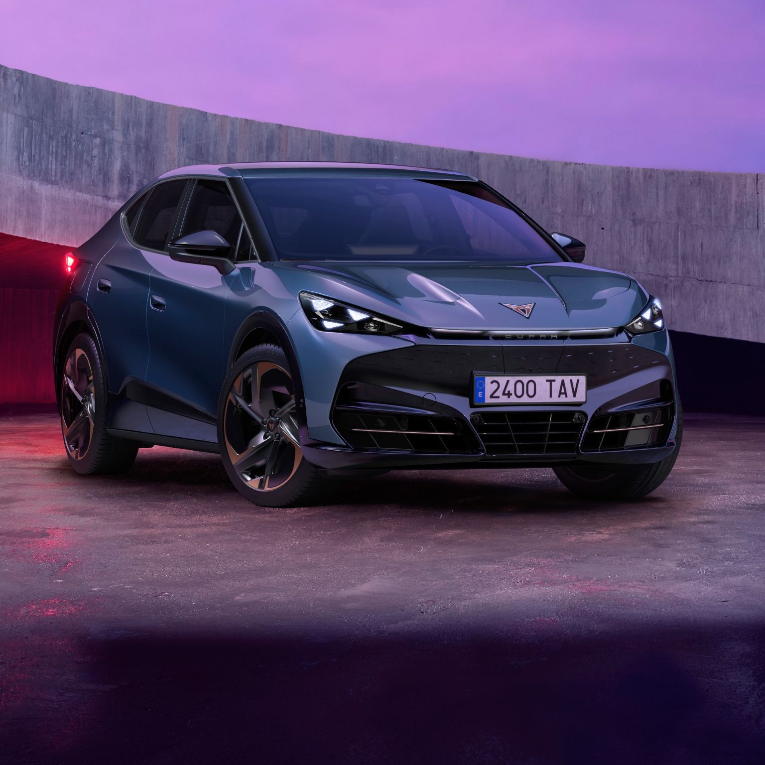 Električni CUPRA TAVASCAN VZ modernega videza, parkiran na betonski površini ob vijoličnemu nebu v ozadju. Avtomobil je modre barve s črnimi detajli. Okolica je osvetljena z mehko svetlobo, ki poudarja linije vozila.