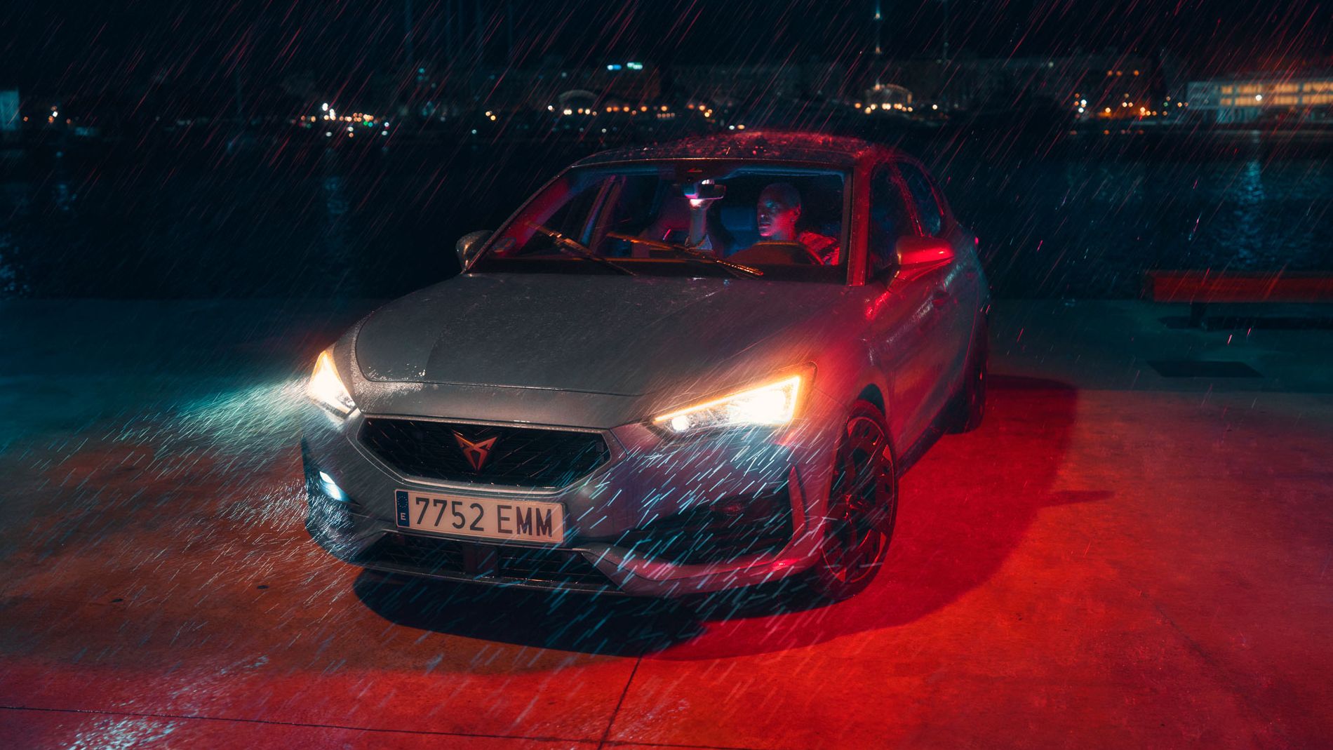 Avtomobil CUPRA Leon 5D z žarometi prižganimi med deževno nočjo. Avto stoji na mokrem asfaltu ob obali, v ozadju so vidne luči mesta. Voznik nastavlja sredinsko vzvratno ogledalo.