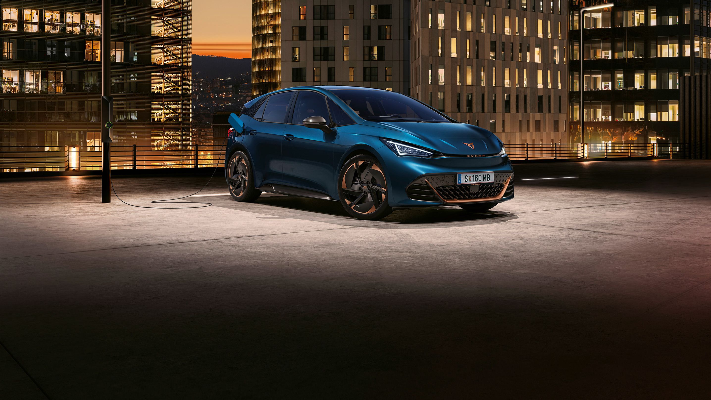 Električni avto CUPRA Born je parkiran in priključen na polnjenje na strehi garažne hiše v mestu ob sončnem zahodu. V ozadju so visoke poslovne stavbe z osvetljenimi okni. 