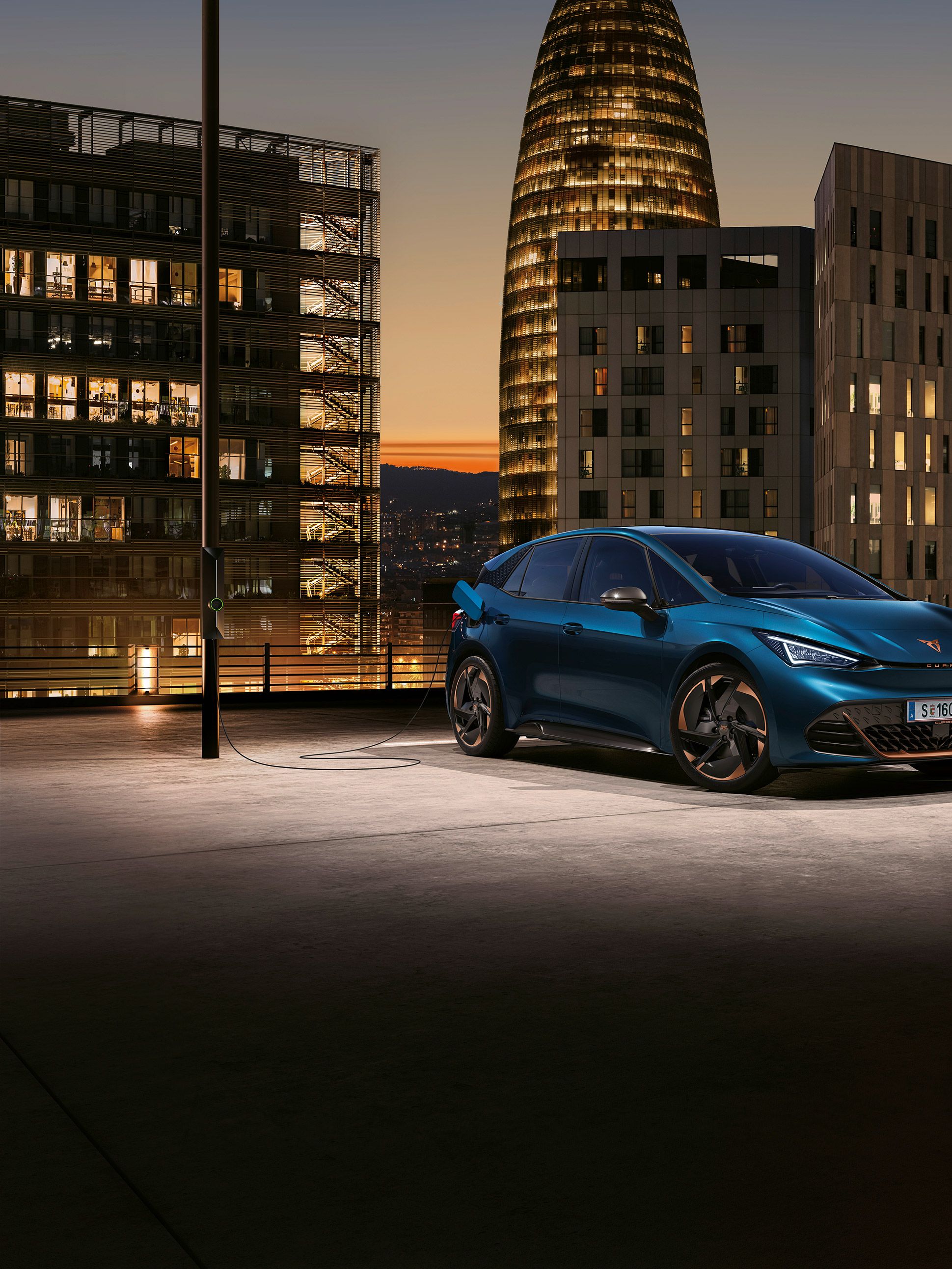 Električni avto CUPRA Born je parkiran in priključen na polnjenje na strehi garažne hiše v mestu ob sončnem zahodu. V ozadju so visoke poslovne stavbe z osvetljenimi okni. 