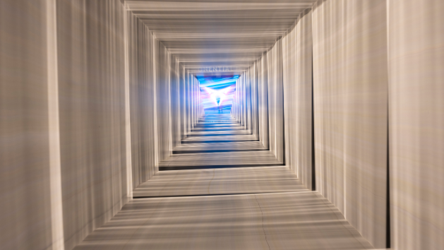 Pogled skozi kvadratni tunel, ki je osvetljen z modro in belo svetlobo. Na koncu tunela je logotip znamke CUPRA. Geometrijske linije ustvarjajo občutek globine in perspektive.