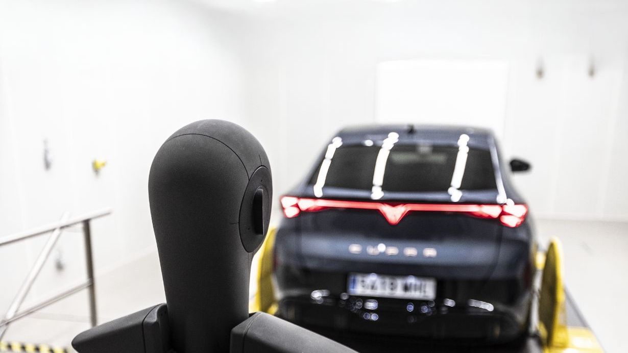 Podoba manekena v testnem laboratoriju opazuje zadnji del temnega avtomobila, ki je nameščen na testni postelji. Avto ima vklopljene zadnje luči. Prostor je dobro osvetljen z belimi stropnimi lučmi.