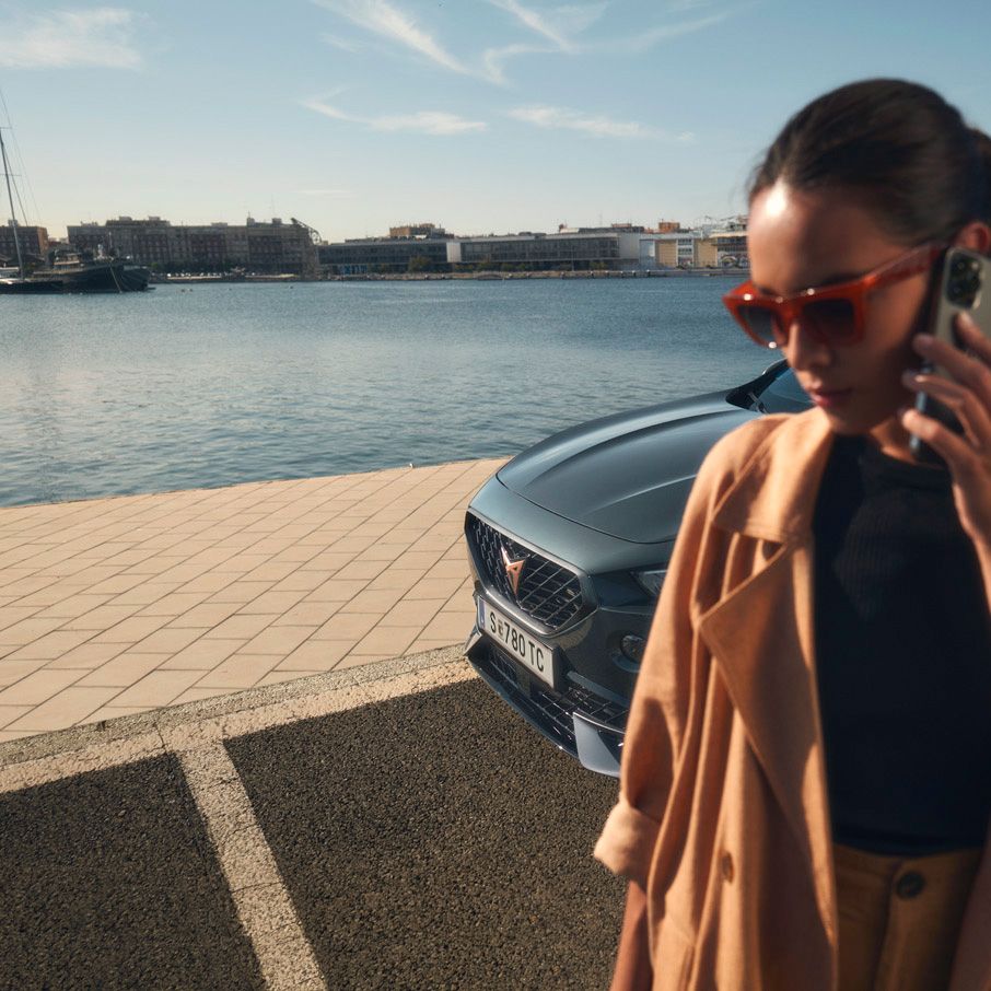 Ženska stoji ob obali in se pogovarja po telefonu. V ozadju je parkiran siv avtomobil CUPRA Formentor. V daljavi so vidne ladje in stavbe ob vodi.