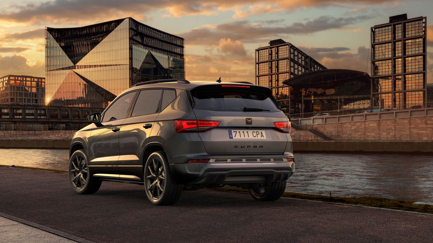 Parkiran CUPRA Ateca ob reki ob sončnem zahodu. V ozadju so sodobne steklene zgradbe in nebo z oblaki. Vozilo je obrnjeno stran od kamere proti reki.