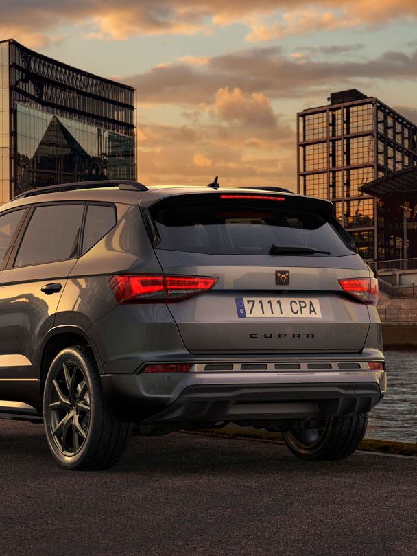 Parkiran CUPRA Ateca ob reki ob sončnem zahodu. V ozadju so sodobne steklene zgradbe in nebo z oblaki. Vozilo je obrnjeno stran od kamere proti reki.