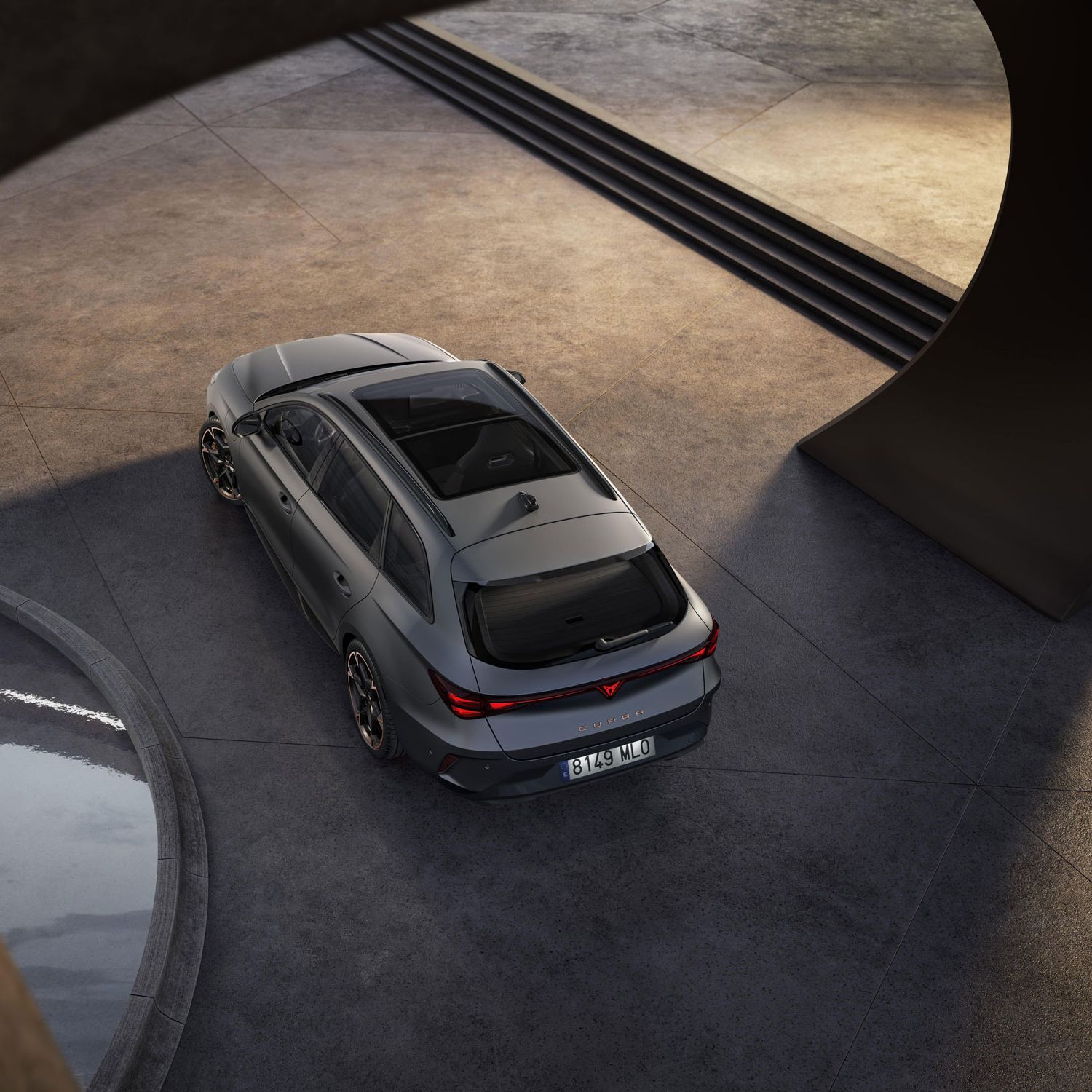 CUPRA LEON SPORTSTOURER VZ parkiran na betonski površini, posnet iz ptičje perspektive. Avto je temno sive barve, okoli njega pa so betonski stebri in majhen bazen z vodo. V ozadju so stopnice, ki vodijo navzgor.