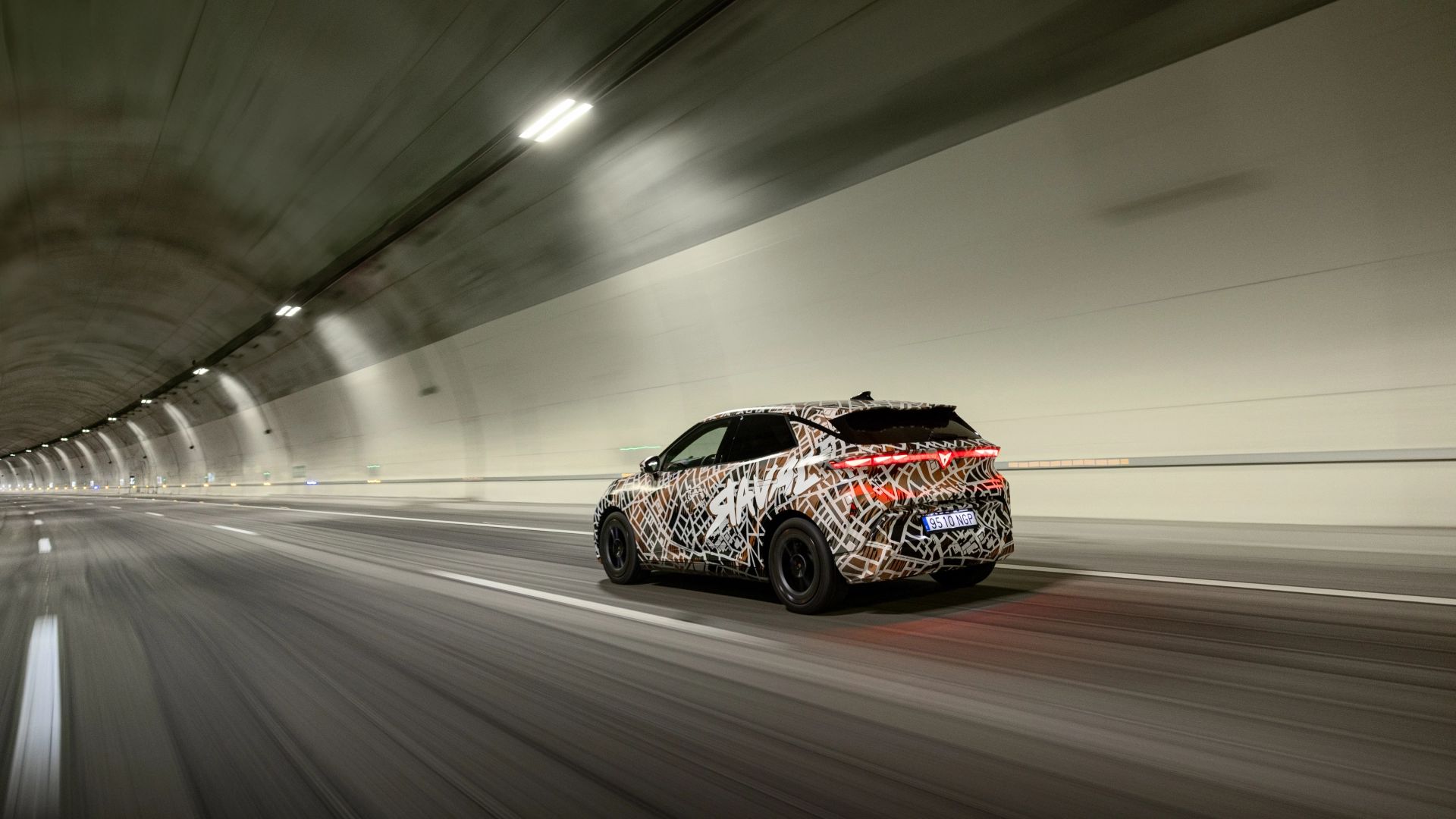 CUPRA Raval Dynamic v maskirni foliji, ki vozi skozi osvetljen tunel. Vozilo je vidno od zadaj. Tunel je osvetljen z lučmi na zgornjem delu sten.