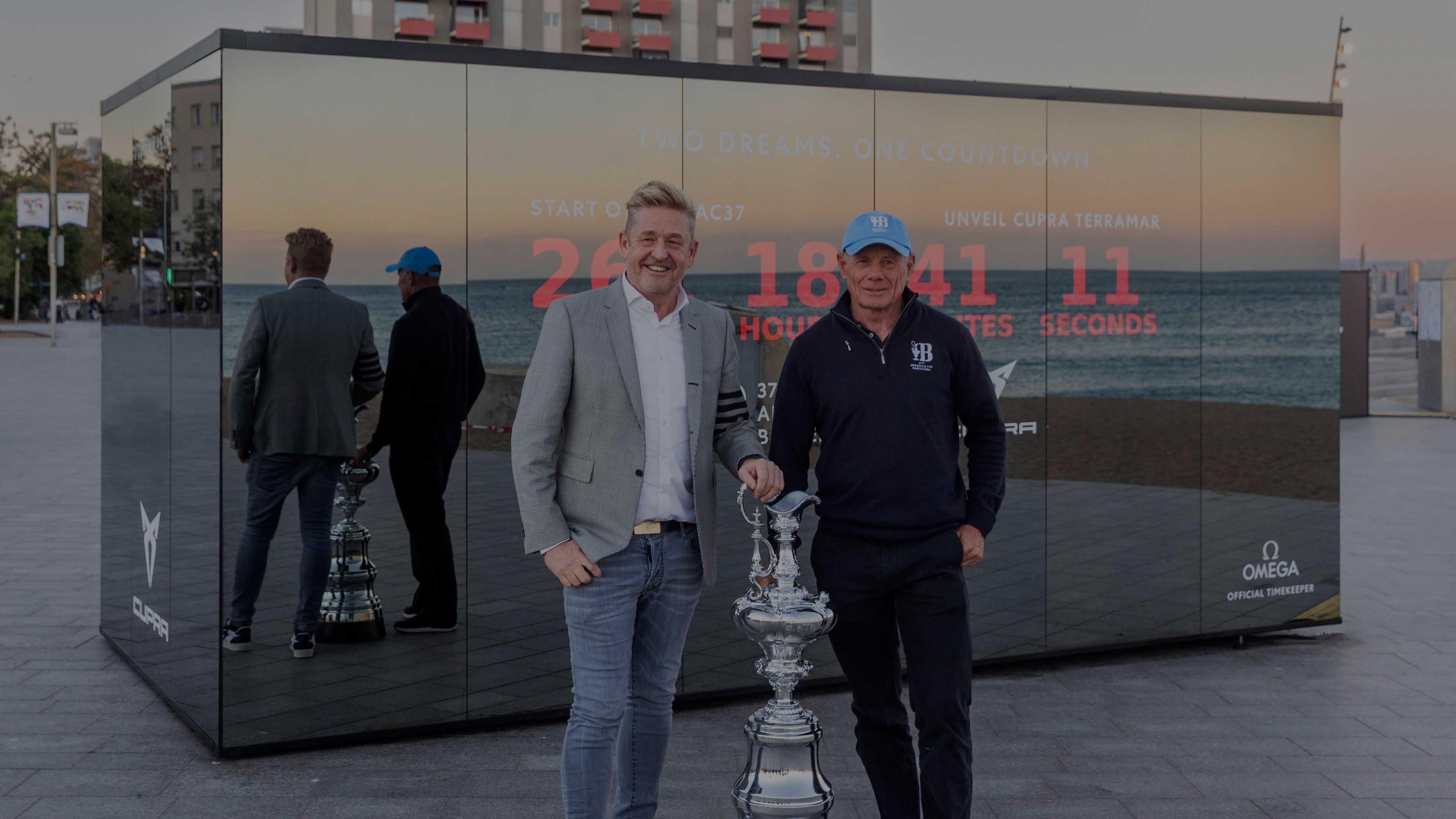 Wayne Griffith in Grant Dalton stojita ob pokalu. Sta na prostem pred veliko stekleno instalacijo, ki prikazuje odštevanje časa in oglaševanje.