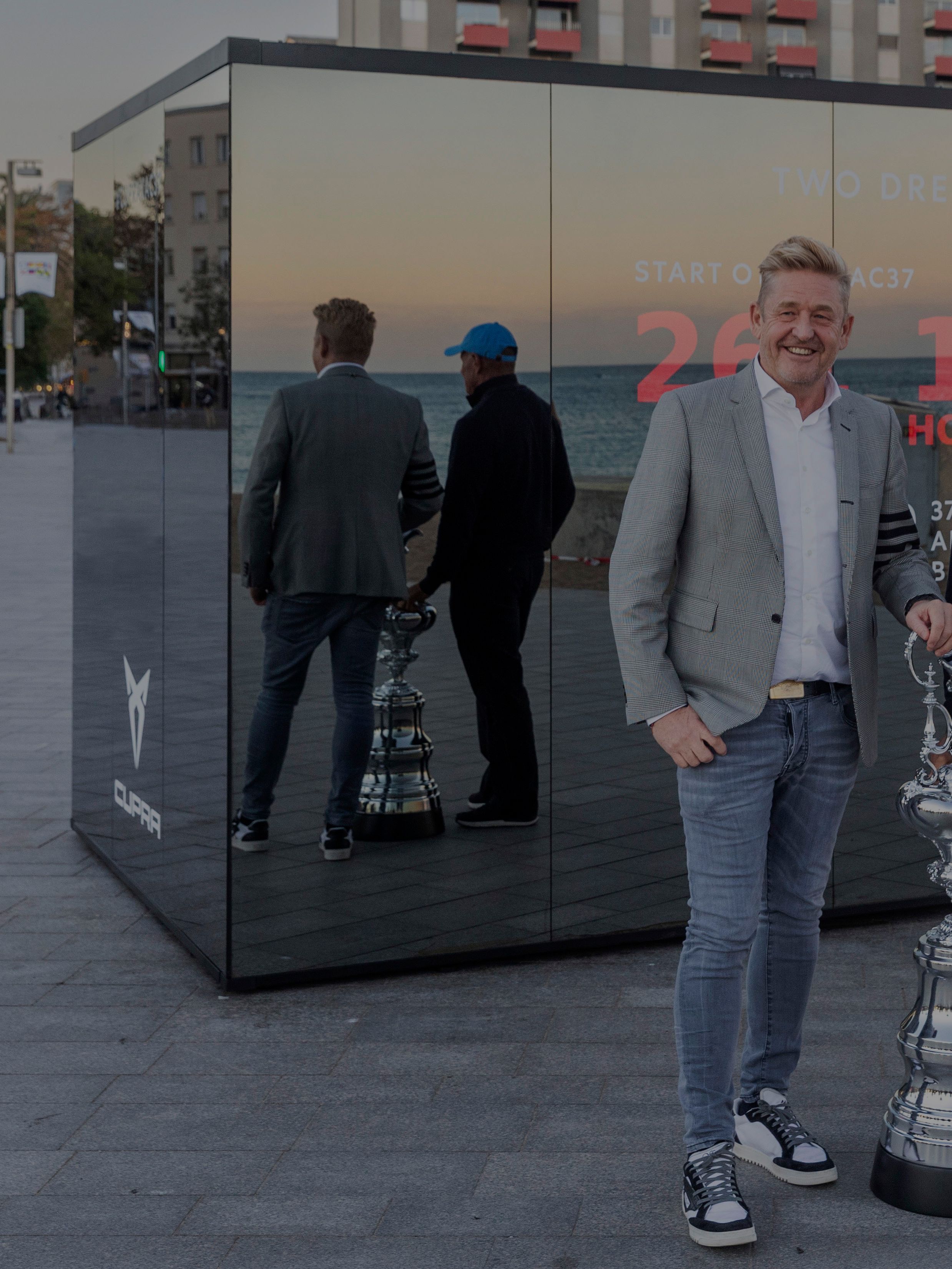 Wayne Griffith in Grant Dalton stojita ob pokalu. Sta na prostem pred veliko stekleno instalacijo, ki prikazuje odštevanje časa in oglaševanje.