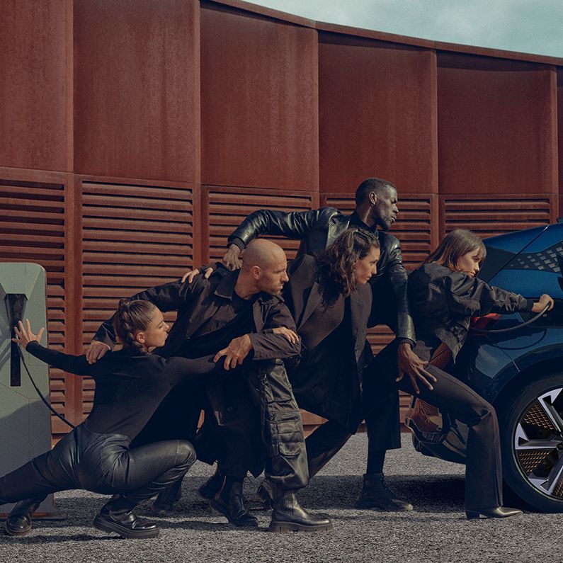Skupina ljudi v črnih oblačilih v različnih pozah, ki se naslanjajo na modro električno vozilo CUPRA Born med polnjenjem. Zraven avtomobila je polnilni post. Scena je dramatična in umetniška.