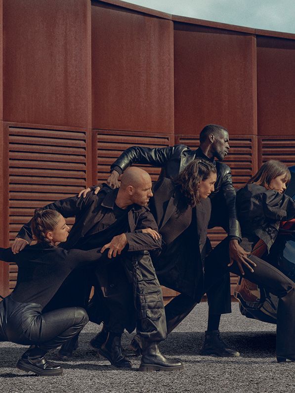 Skupina ljudi v črnih oblačilih v različnih pozah, ki se naslanjajo na modro električno vozilo CUPRA Born med polnjenjem. Zraven avtomobila je polnilni post. Scena je dramatična in umetniška.