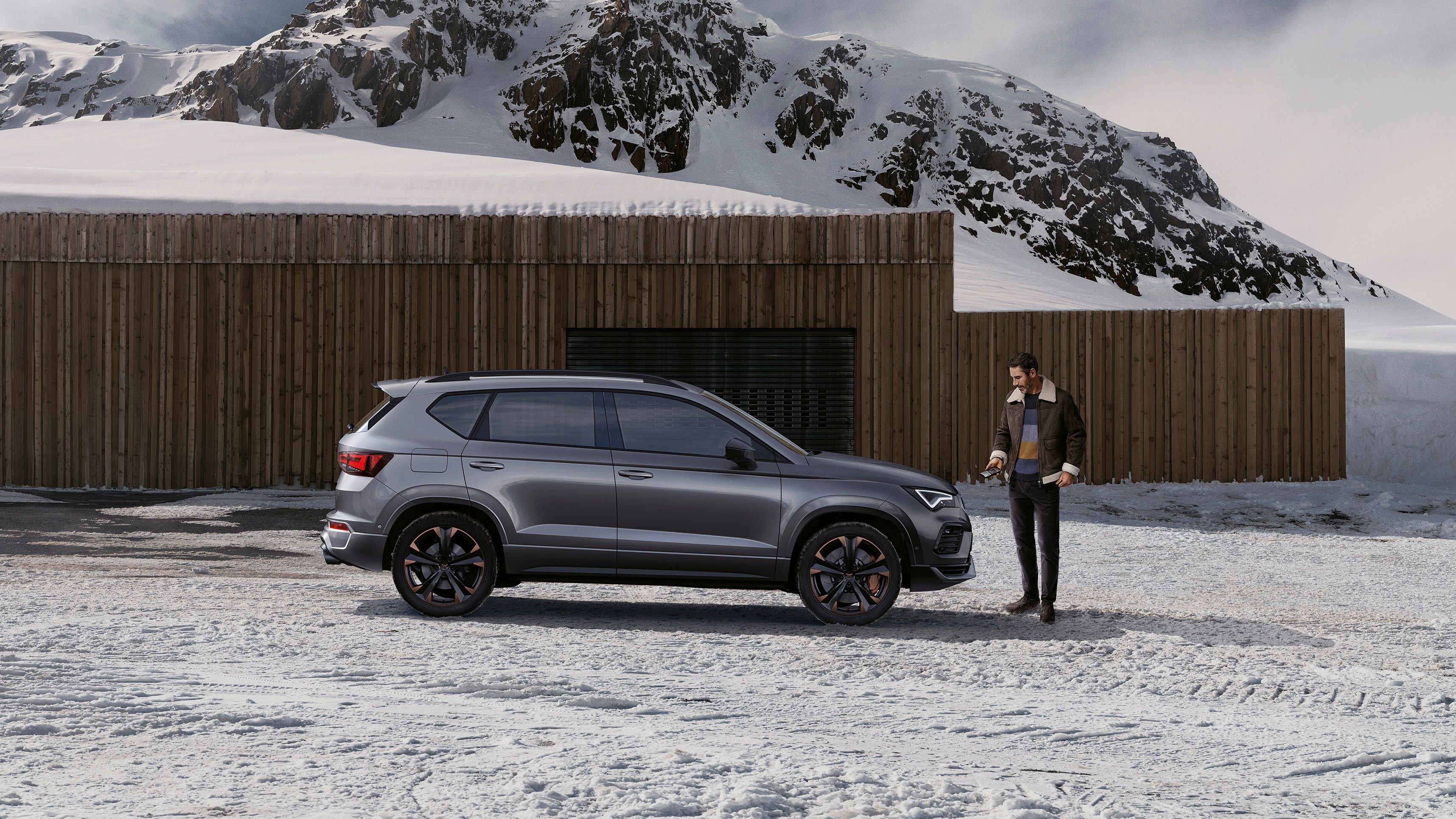 Moški stoji pred sivo vozilo CUPRA Ateca, parkirano pred lesenim objektom v zasneženem gorskem okolju. V ozadju se vidijo visoki, zasneženi vrhovi gora.