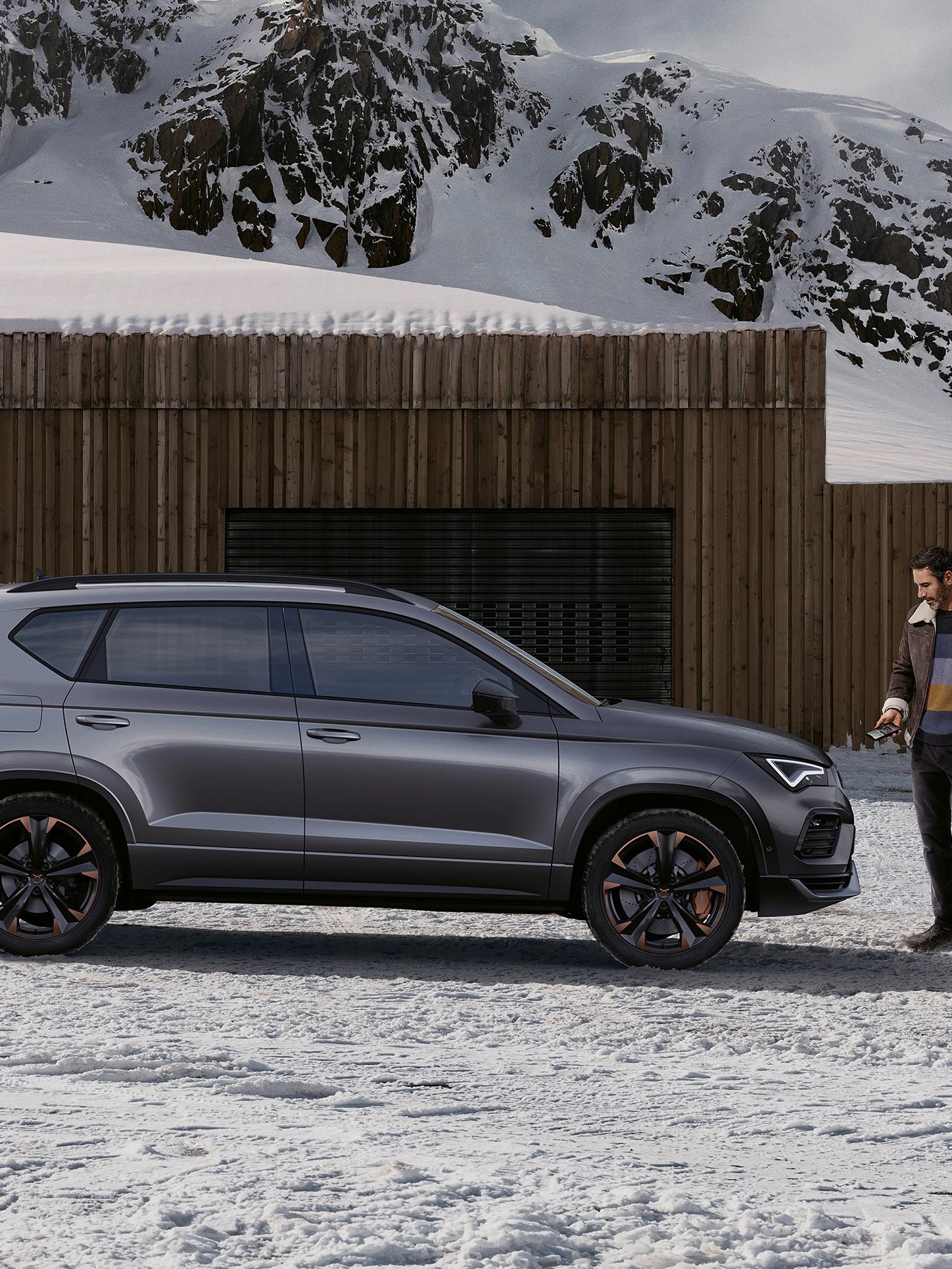 Moški stoji pred sivo vozilo CUPRA Ateca, parkirano pred lesenim objektom v zasneženem gorskem okolju. V ozadju se vidijo visoki, zasneženi vrhovi gora.