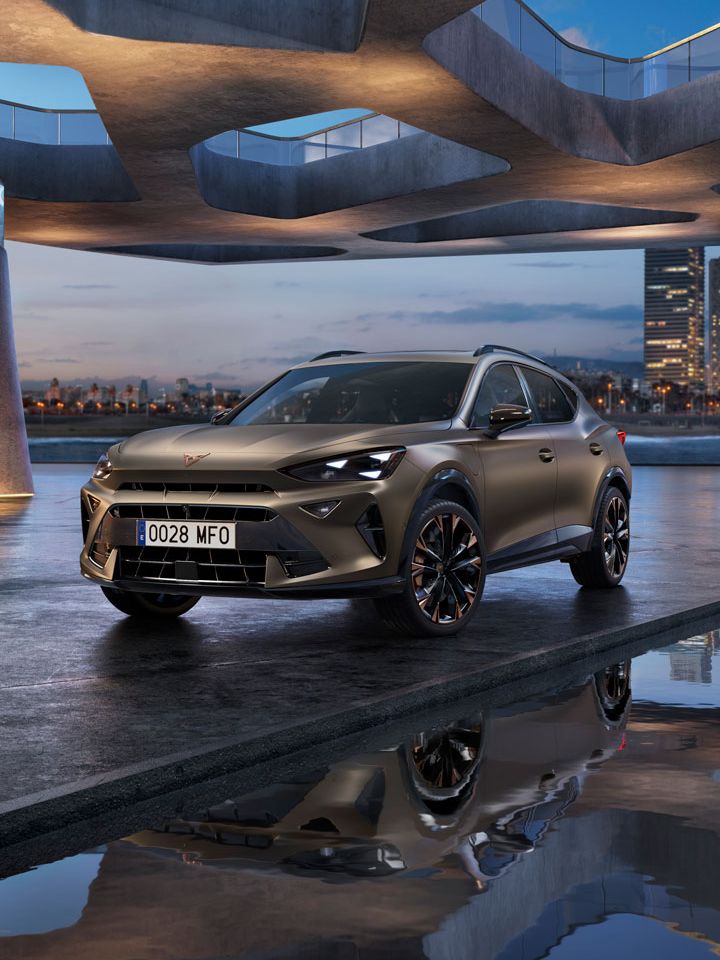 Parkiran CUPRA Formentor PA na zunanjem sodobnem parkirišču z zanimivo arhitekturo. V ozadju se vidi mestna panorama ob reki. Nebo je modro ob sončnem zahodu.