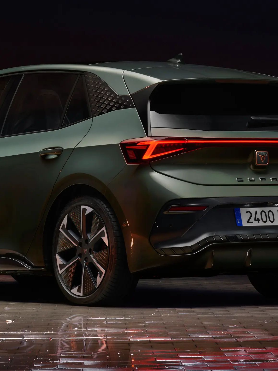 Zadnja stran zelenega Cupra Born avtomobila, parkiranega v urbanem okolju ponoči, z osvetljenimi stavbami v ozadju. Avtomobil ima sodobno oblikovane luči in lita platišča.