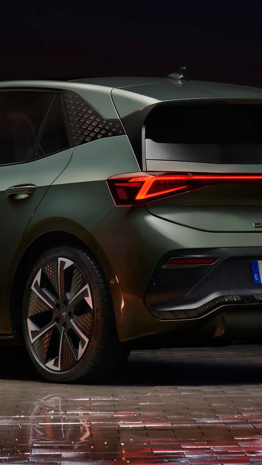 Zadnja stran zelenega Cupra Born avtomobila, parkiranega v urbanem okolju ponoči, z osvetljenimi stavbami v ozadju. Avtomobil ima sodobno oblikovane luči in lita platišča.