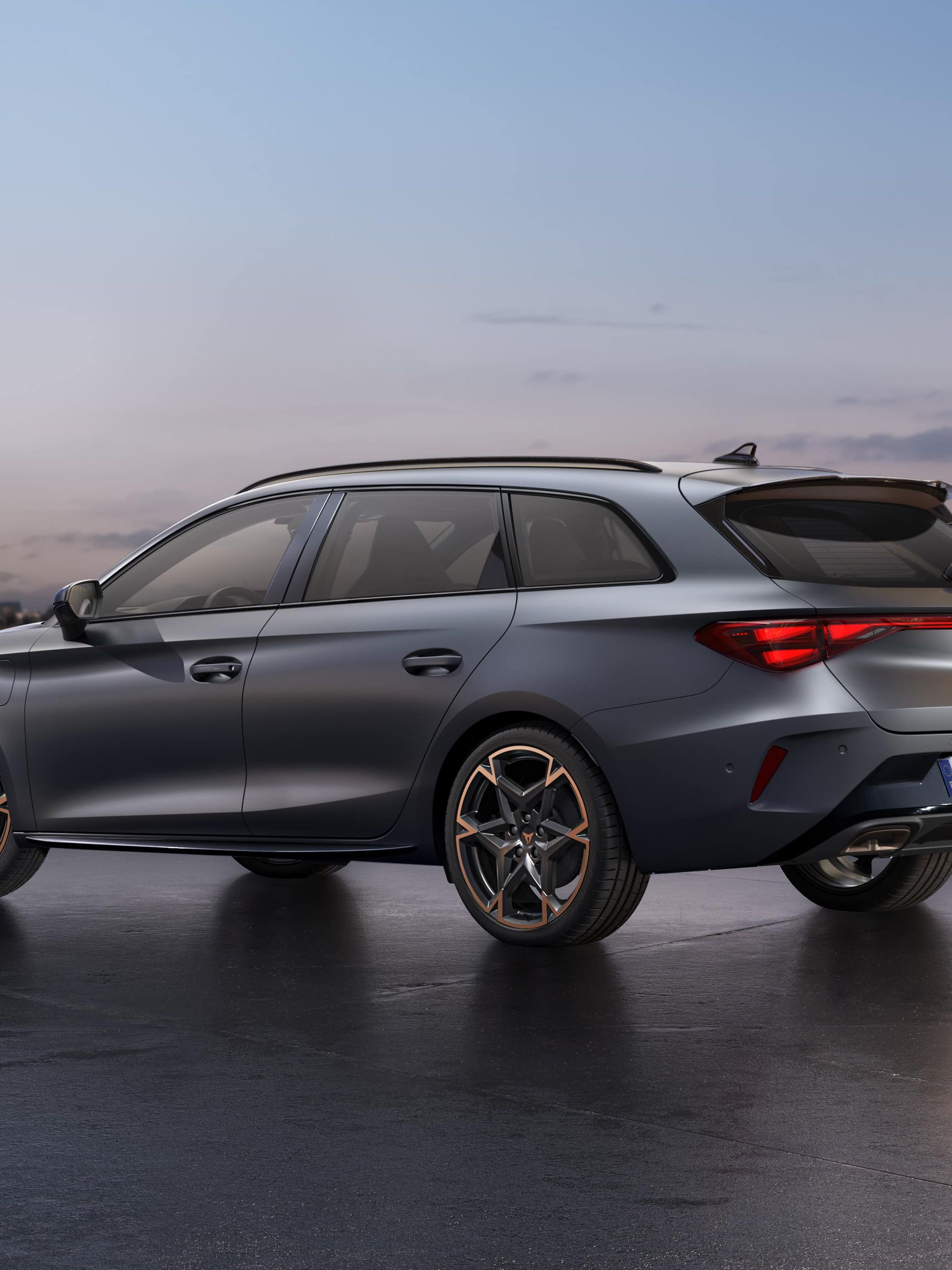 CUPRA LEON SPORTSTOURER stoji ob obali ob sončnem zahodu. V ozadju je mestno obzorje z visokimi stavbami, mokra površina pod vozilom pa odseva nebo.