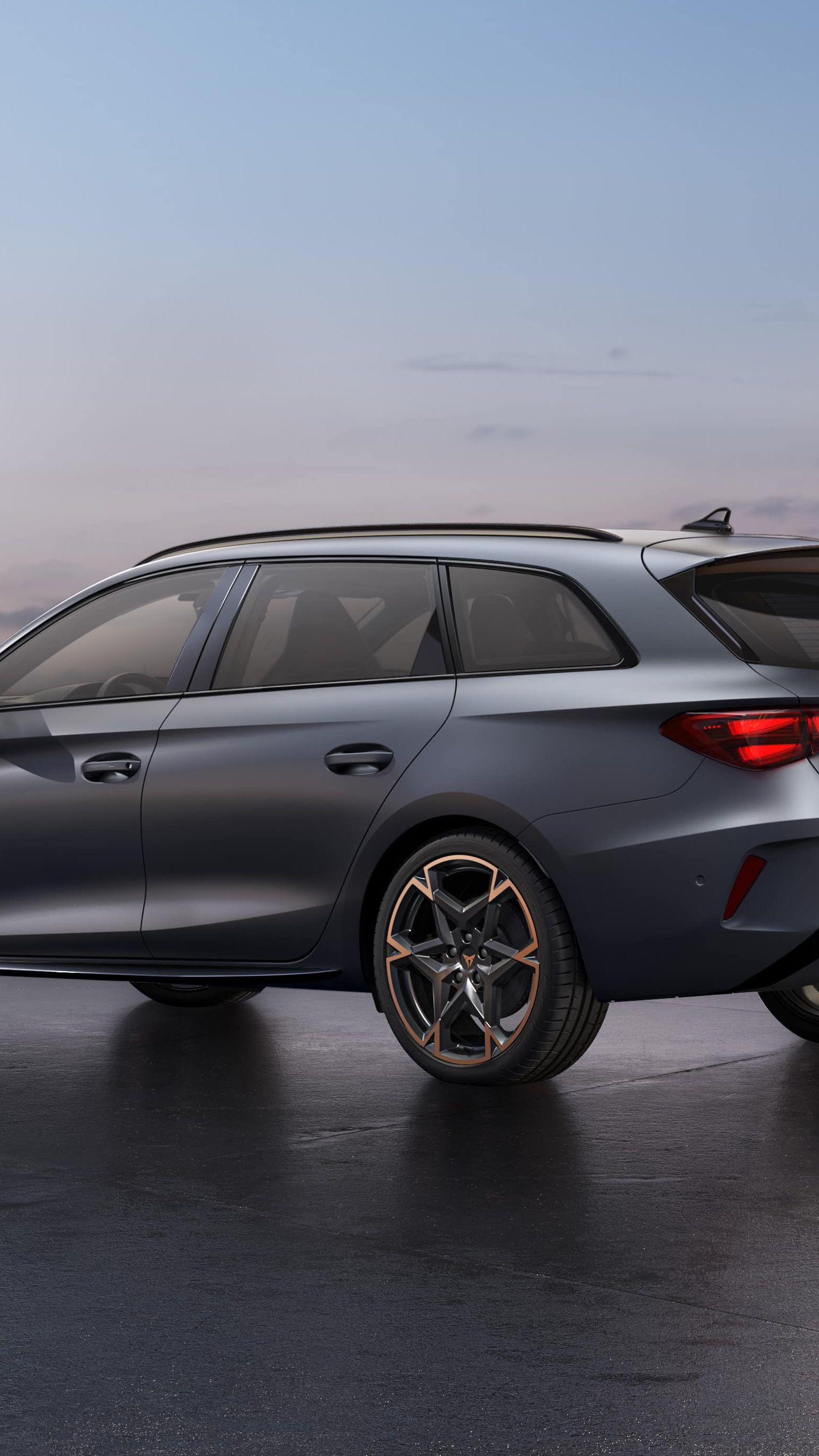 CUPRA LEON SPORTSTOURER stoji ob obali ob sončnem zahodu. V ozadju je mestno obzorje z visokimi stavbami, mokra površina pod vozilom pa odseva nebo.