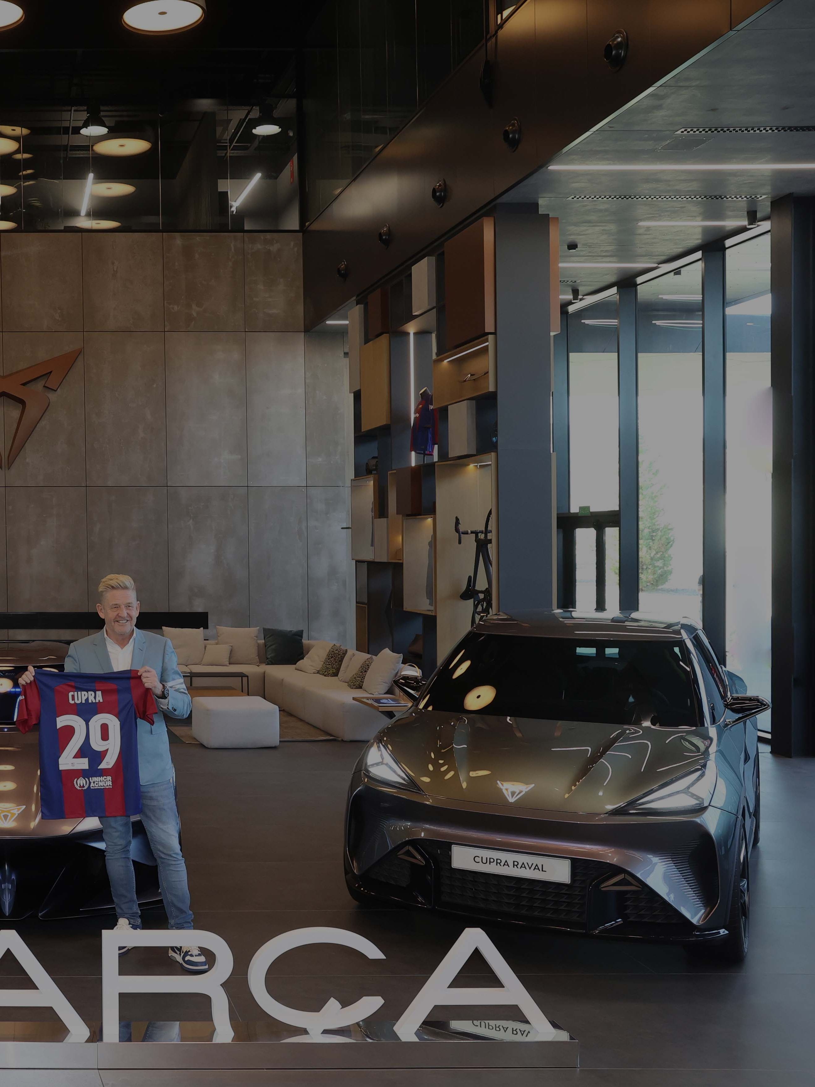 Dva moška držita majici nogometnega kluba pred razstavljenimi CUPRA avtomobili, CUPRA Tavascan in CUPRA Raval v moderno opremljenem salonu. Na tabli pred njima je napis "I LOVE BARÇA".