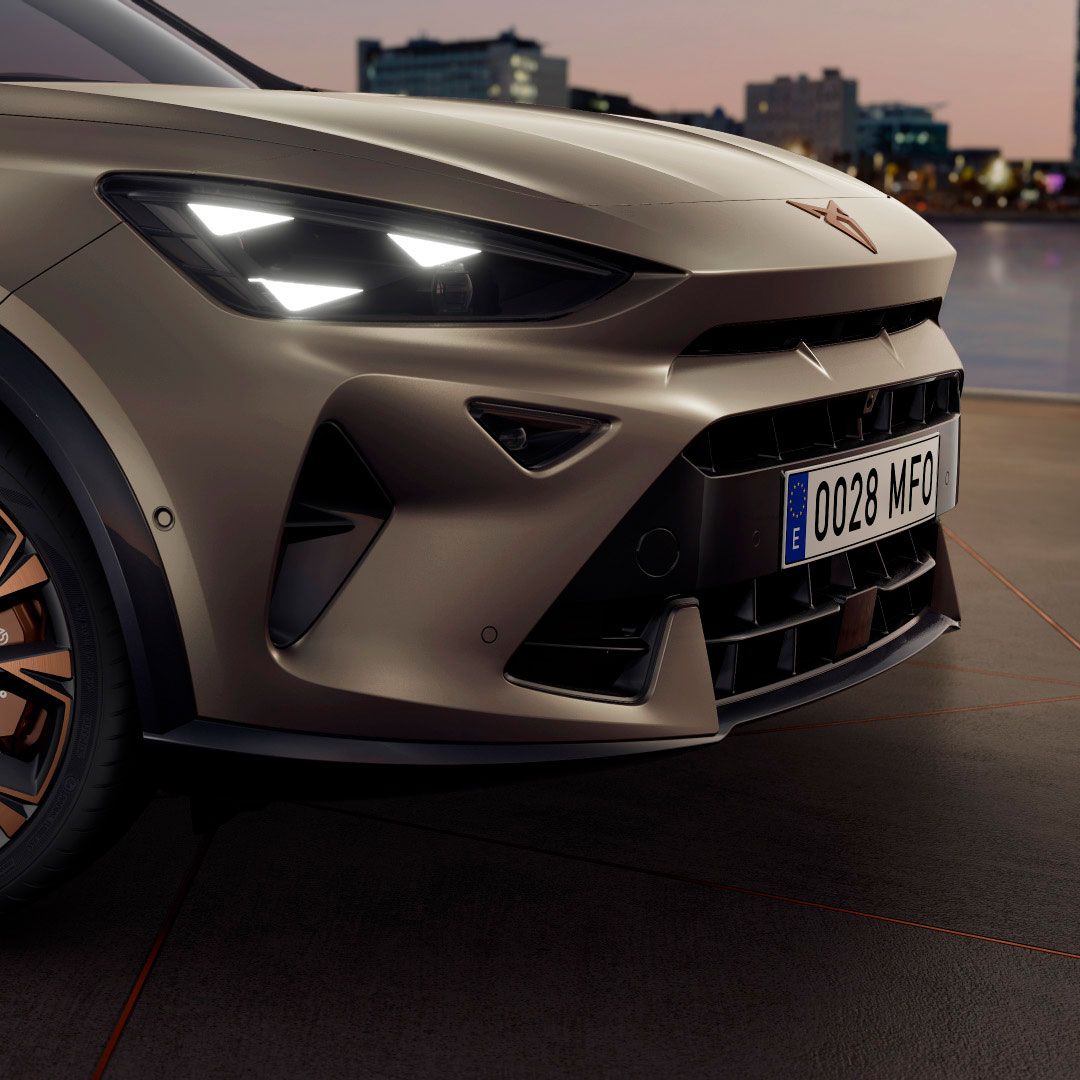 CUPRA Formentor PA z vključenimi sprednjimi žarometi na ploščadi ob vodi ob sončnem zahodu. Avto ima mat zlato-rjavo barvo ter črno-oranžna platišča. V ozadju je mestna obala.