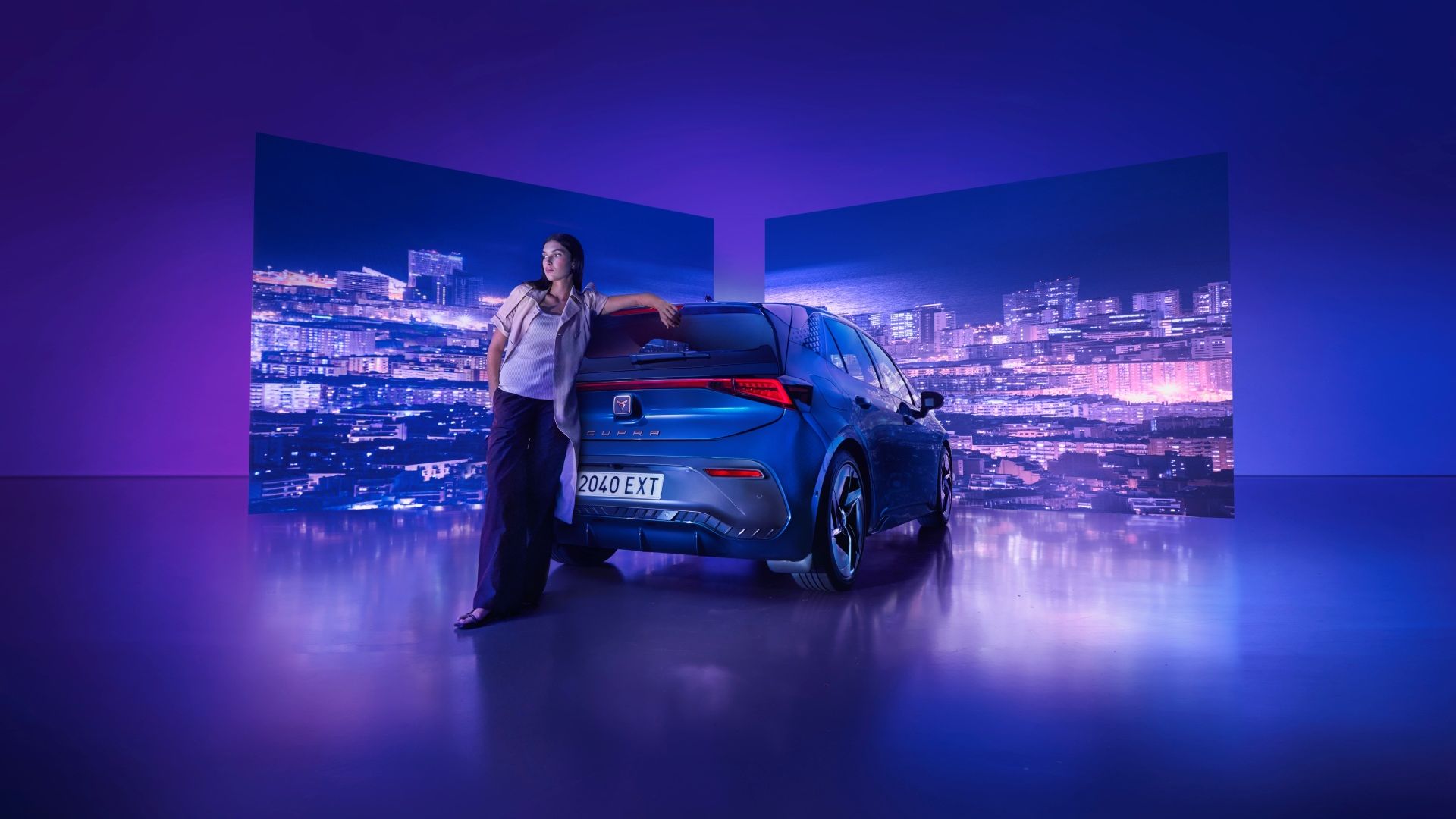 Ženska, stoji zraven parkiranega modrega avtomobila CUPRA Born v osvetljenem prostoru, z mestnim nebom na dva ekrana v ozadju. Ozadje je obarvano v modrih in vijoličnih tonih.