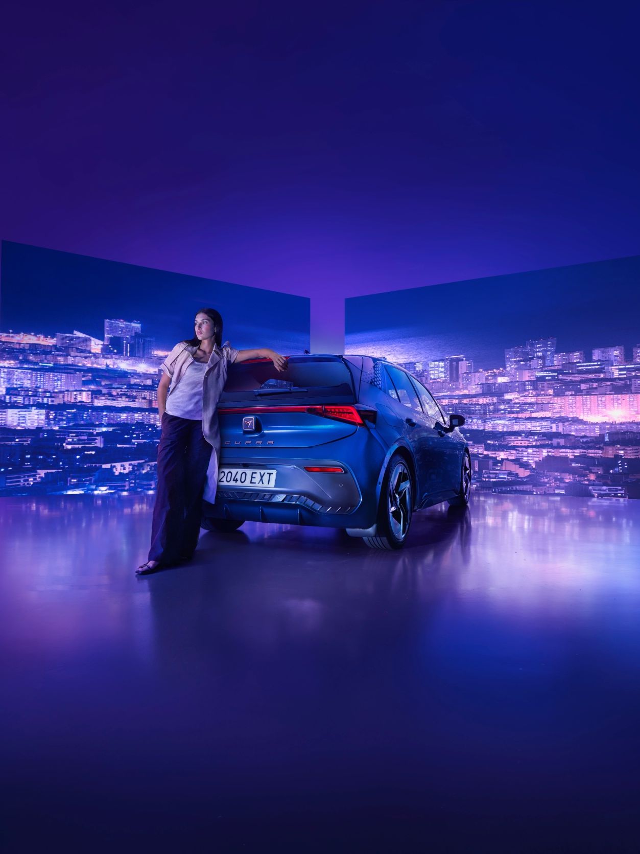 Ženska, stoji zraven parkiranega modrega avtomobila CUPRA Born v osvetljenem prostoru, z mestnim nebom na dva ekrana v ozadju. Ozadje je obarvano v modrih in vijoličnih tonih.