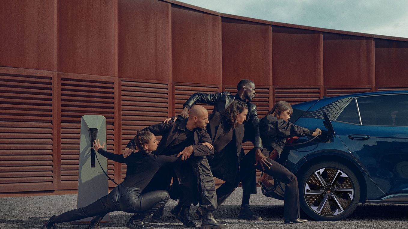 Skupina ljudi v črnih oblačilih v različnih pozah, ki se naslanjajo na modro električno vozilo CUPRA Born med polnjenjem. Zraven avtomobila je polnilni post. Scena je dramatična in umetniška.