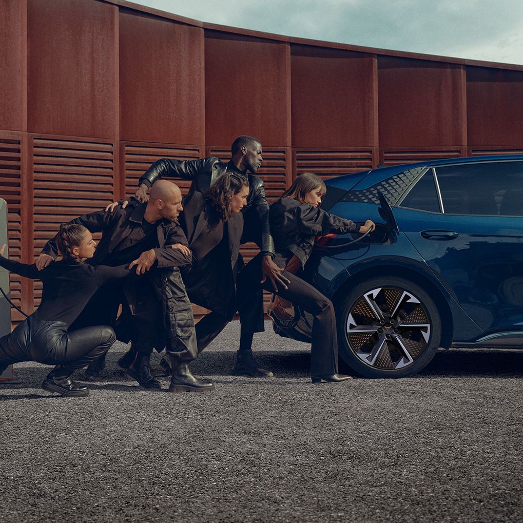 Skupina ljudi v črnih oblačilih v različnih pozah, ki se naslanjajo na modro električno vozilo CUPRA Born med polnjenjem. Zraven avtomobila je polnilni post. Scena je dramatična in umetniška.