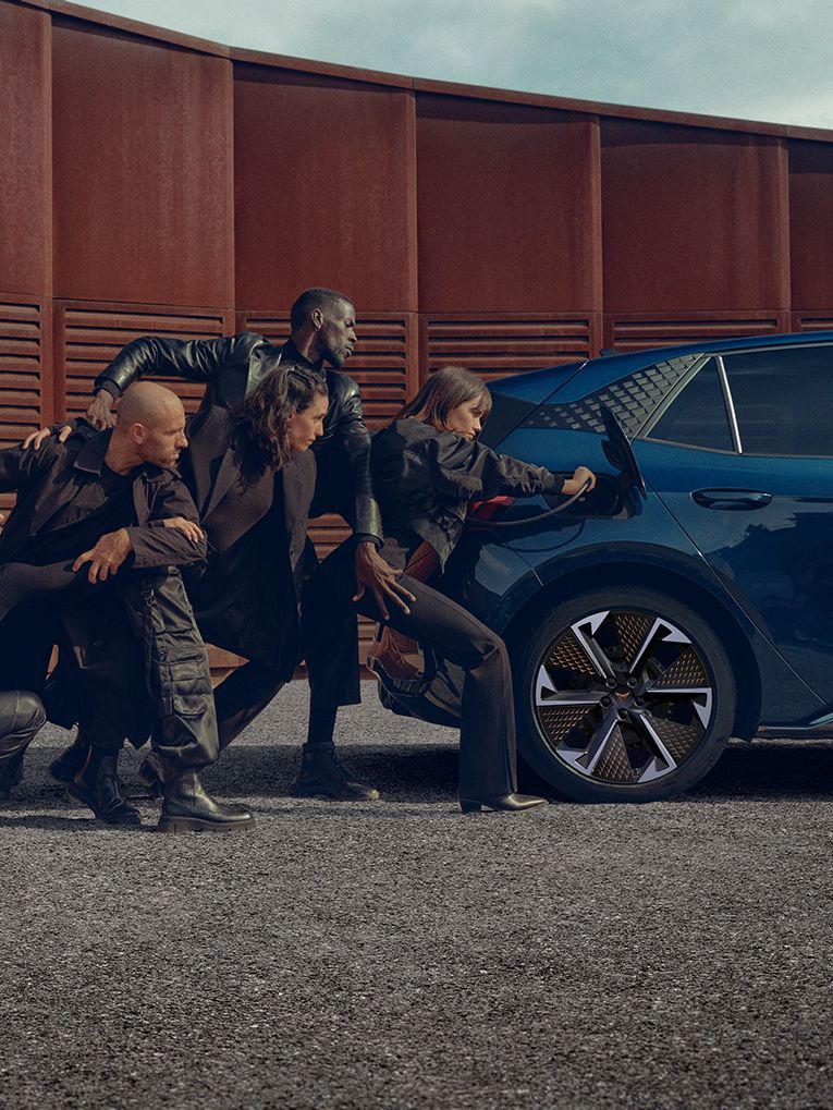Skupina ljudi v črnih oblačilih v različnih pozah, ki se naslanjajo na modro električno vozilo CUPRA Born med polnjenjem. Zraven avtomobila je polnilni post. Scena je dramatična in umetniška.