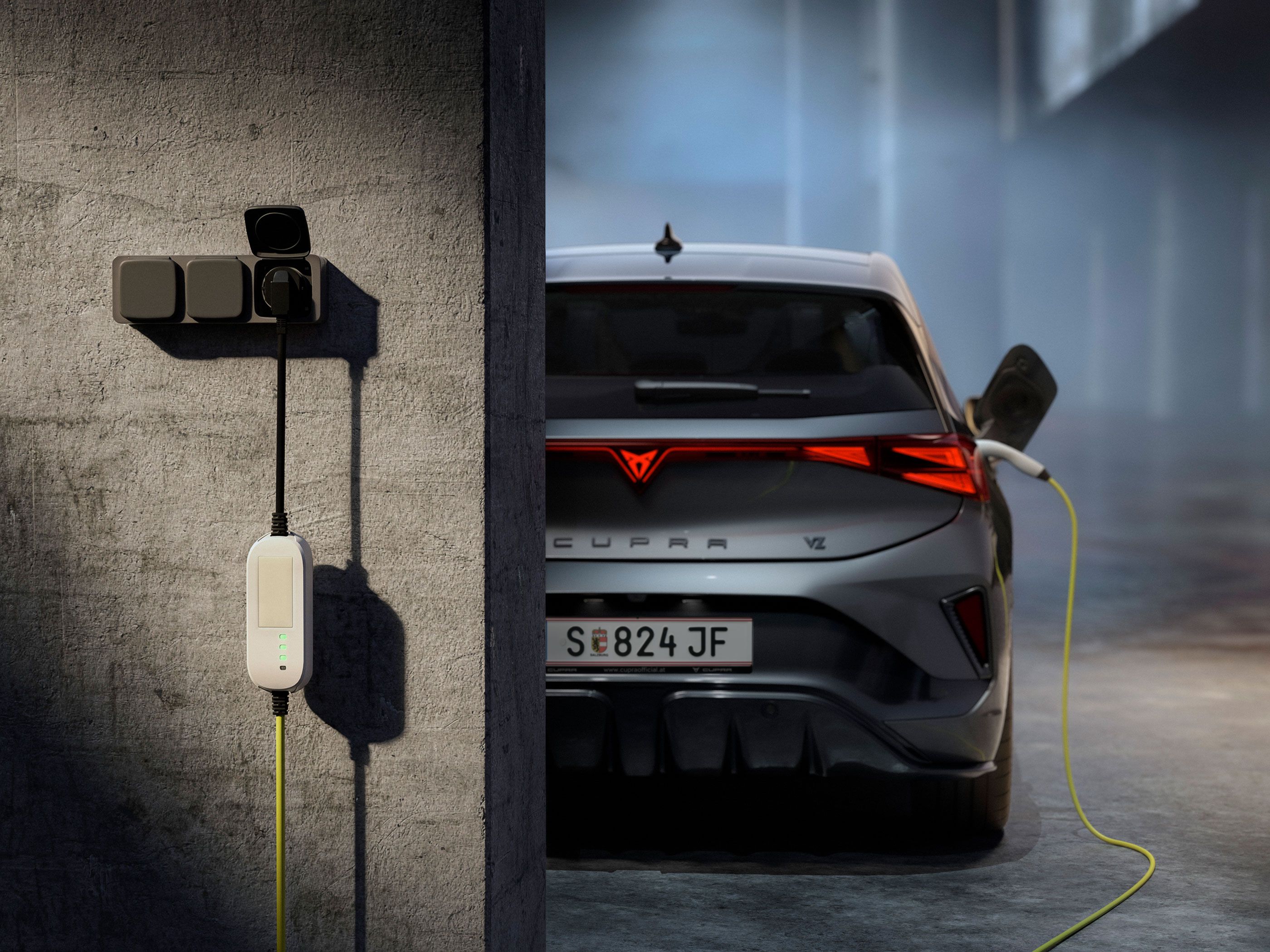 Električni avto CUPRA BORN je parkiran v garaži in priključen na stensko polnilno postajo z rumenim kablom. Leva stran slike prikazuje siv betonski zid z nameščenim polnilnikom, desna pa zadnji del avtomobila s prižganimi zadnjimi lučmi. V ozadju je zamegljen, industrijsko videti prostor.