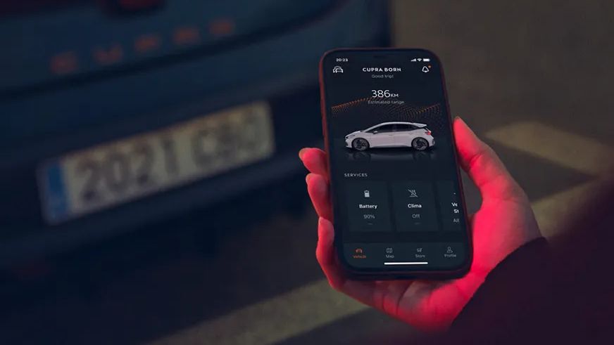 Oseba drži pametni telefon in uporablja 'My CUPRA app'. V ozadju je zadnji del avtomobila z osvetljenimi rdečimi lučmi. 