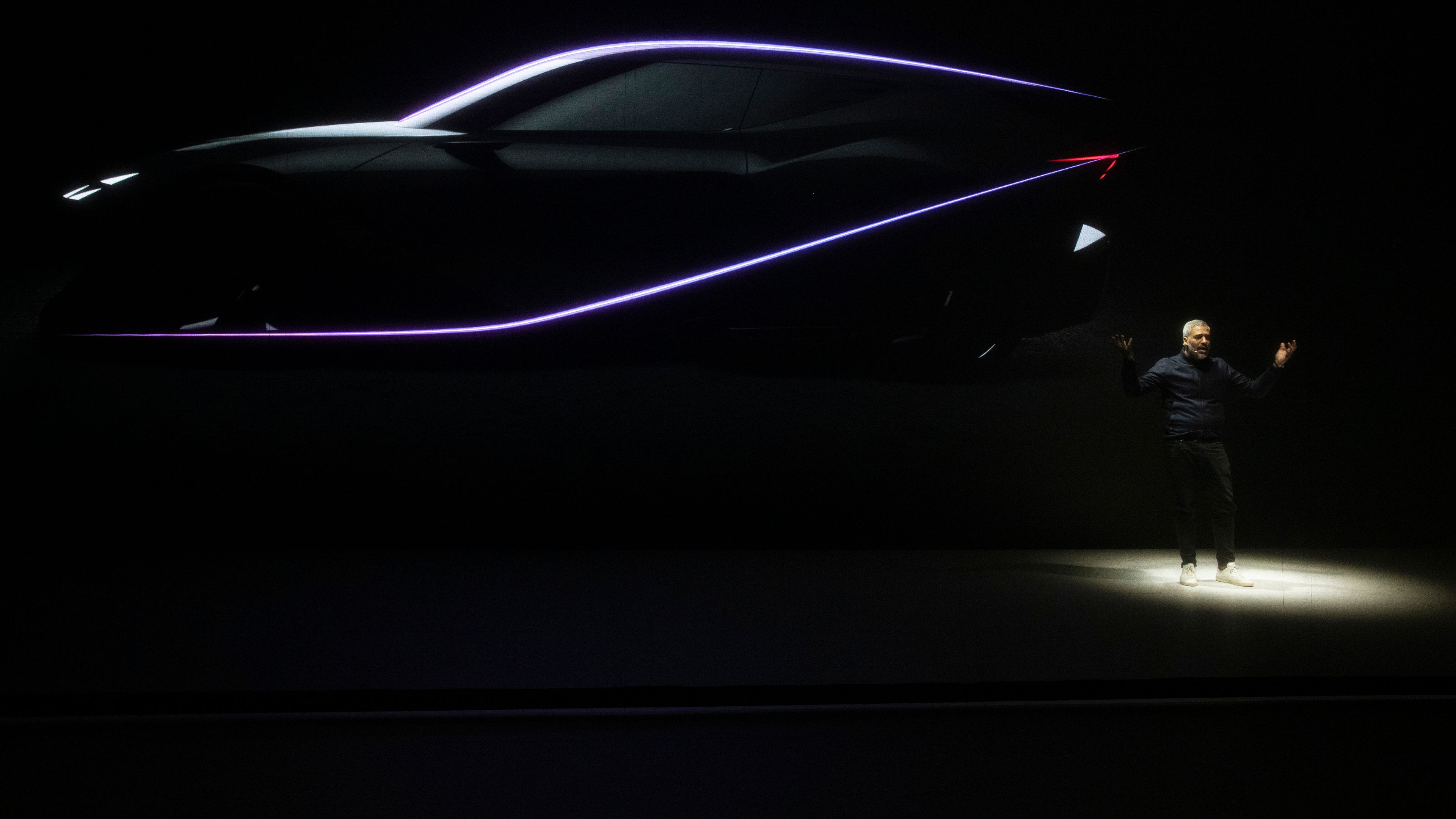 Na odru je temen futurističen avtomobil CUPRA DarkRebel, ki je osvetljen s svetlečo linijo. Oblikovalec Jorge Díez stoji pod njim in gestikulira z rokami.