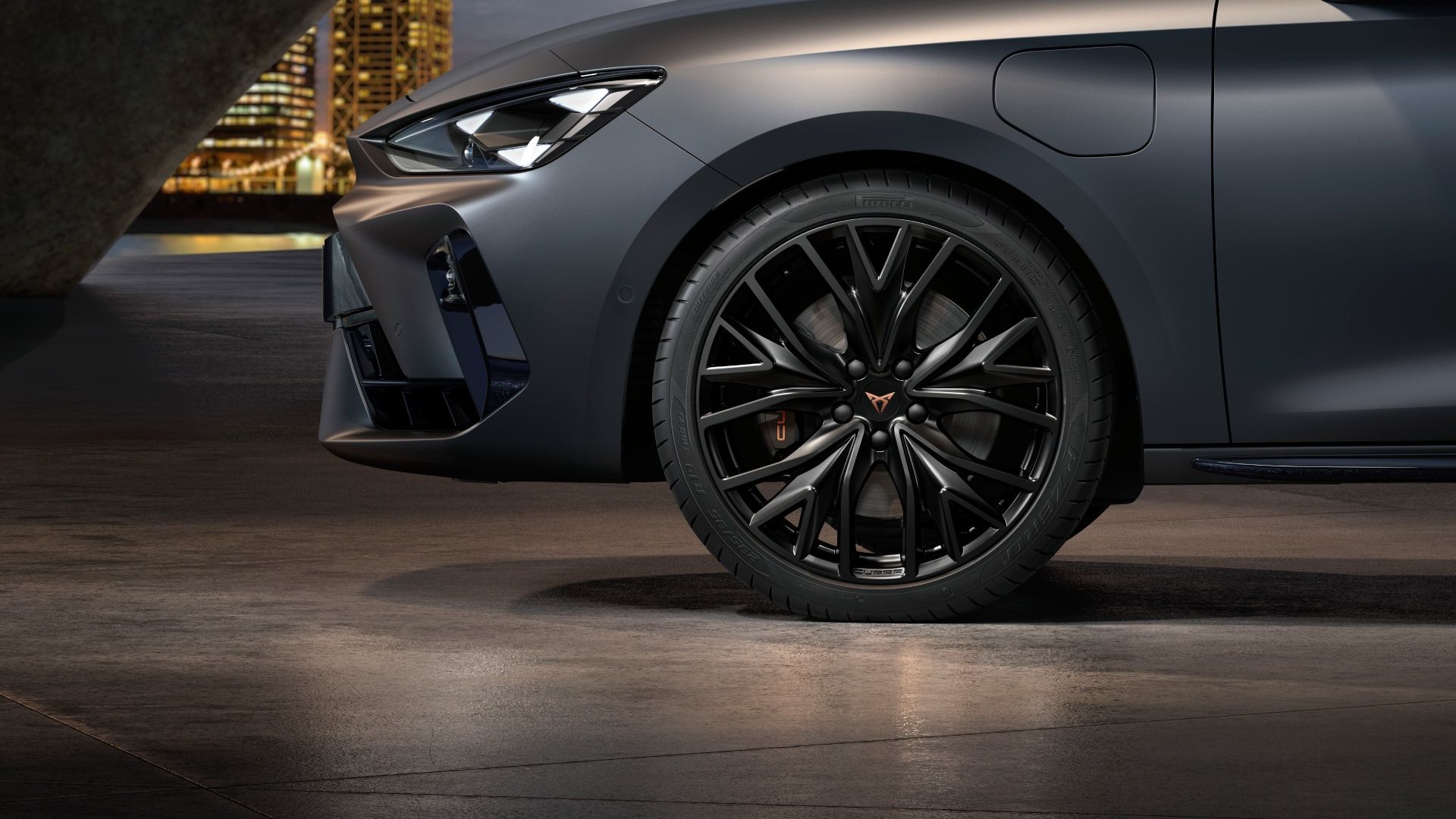 Na sliki je stranski pogled na sprednje kolo in sprednji del mat temno sivega avtomobila CUPRA Leon. Kolo ima športna črna platišča. Avtomobil je parkiran v  sodobi garaži. V ozadju so osvetljene mestne stavbe.