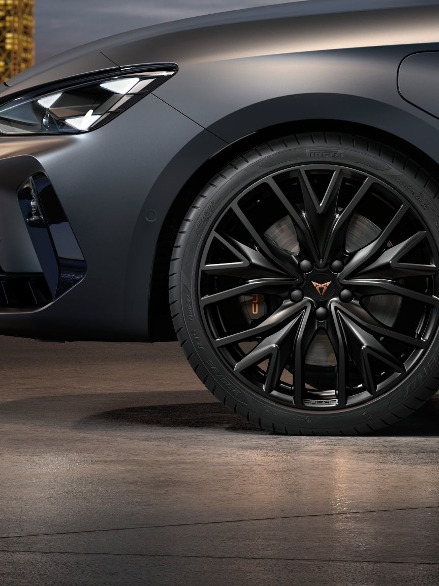Na sliki je stranski pogled na sprednje kolo in sprednji del mat temno sivega avtomobila CUPRA Leon. Kolo ima športna črna platišča. Avtomobil je parkiran v  sodobi garaži. V ozadju so osvetljene mestne stavbe.