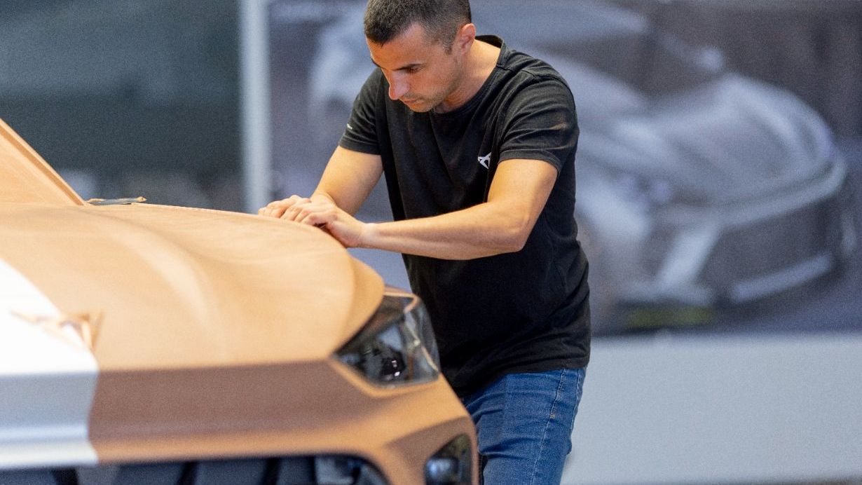 Moški v črni majici stoji ob maketi avtomobila znamke CUPRA iz gline in z rokami natančno oblikuje sprednji del vozila v oblikovalskem studiu.