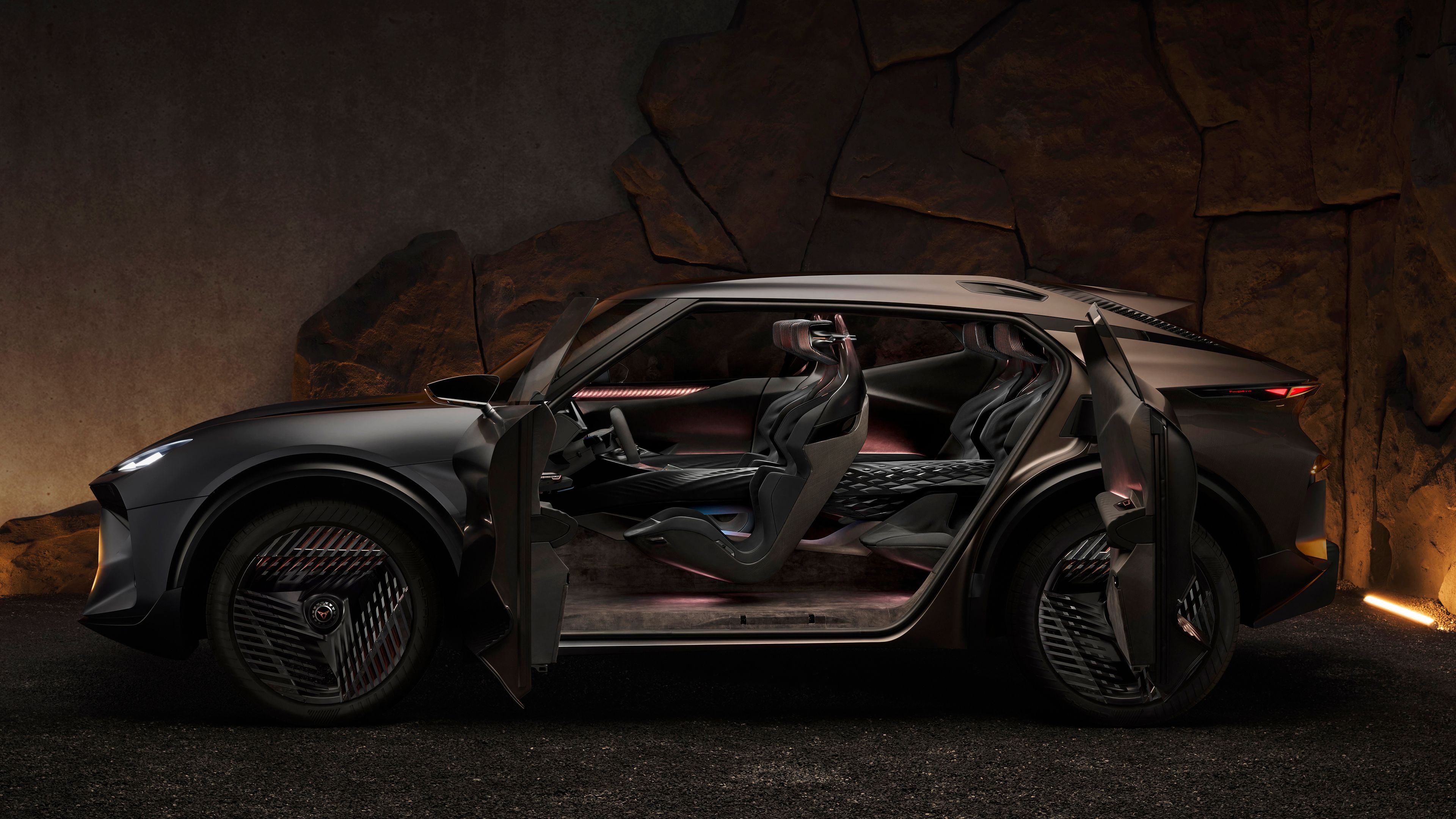 Stranski pogled na luksuzno vozilo CUPRA Tindaya z odprtimi vrati, ki razkrivajo moderno, osvetljeno notranjost. V ozadju je kamnita stena, ki ustvarja kontrast z elegantnim dizajnom vozila.