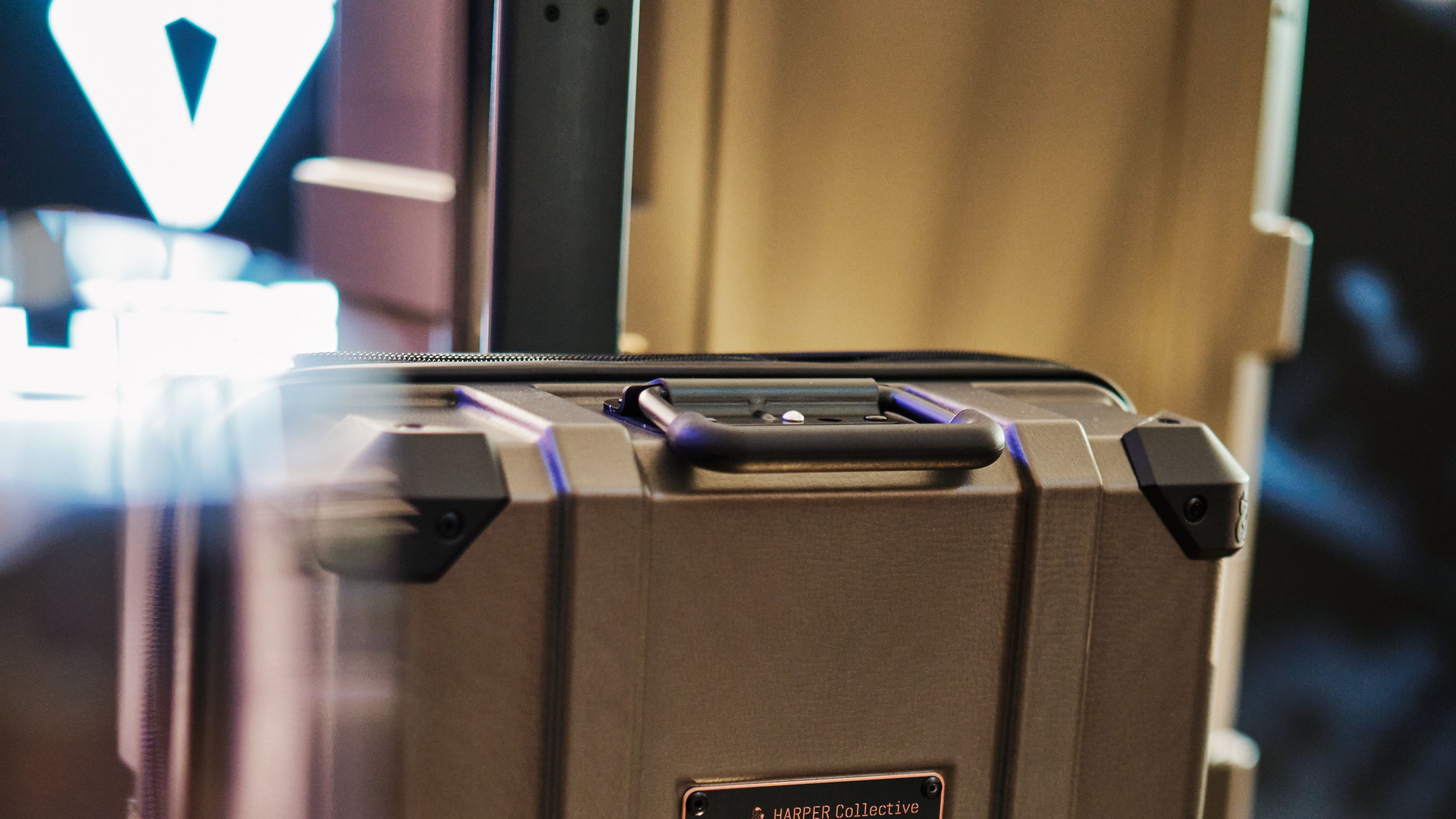 Na sliki je robusten kovček s kolesi, ki je postavljen na mrežasto površino. Kovček ima pravokotno ploščico, ki vključuje logotip znamke CUPRA. Ozadje je zamegljeno in prikazuje notranjost trgovine.