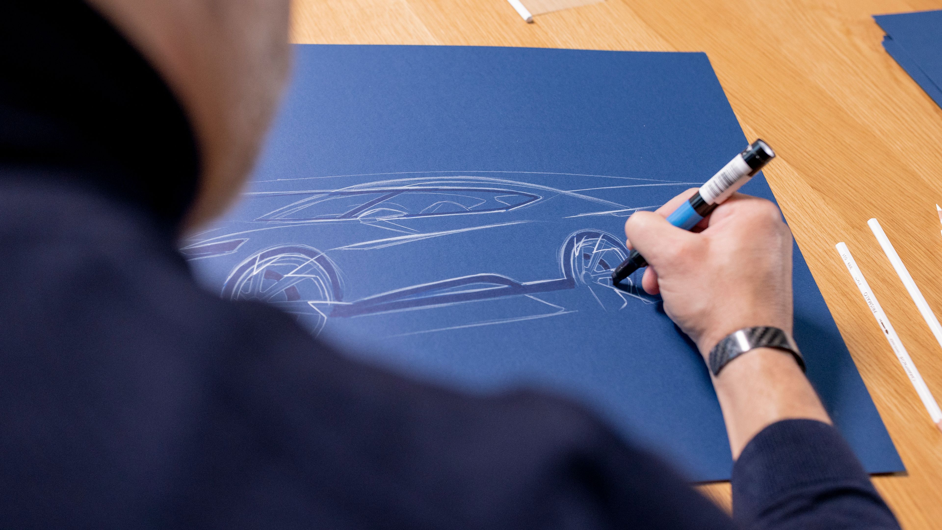 Oblikovalec riše skico avtomobila znamke CUPRA na temno modrem papirju. Skica prikazuje profil športnega avtomobila z poudarkom na kolesih in aerodinamičnih linijah.
