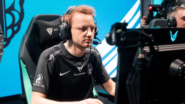 Moški sedi pred računalnikom in igra video igro na tekmovanju. Ima slušalke in mikrofon. V ozadju je zaslon z modro-sivim vzorcem.