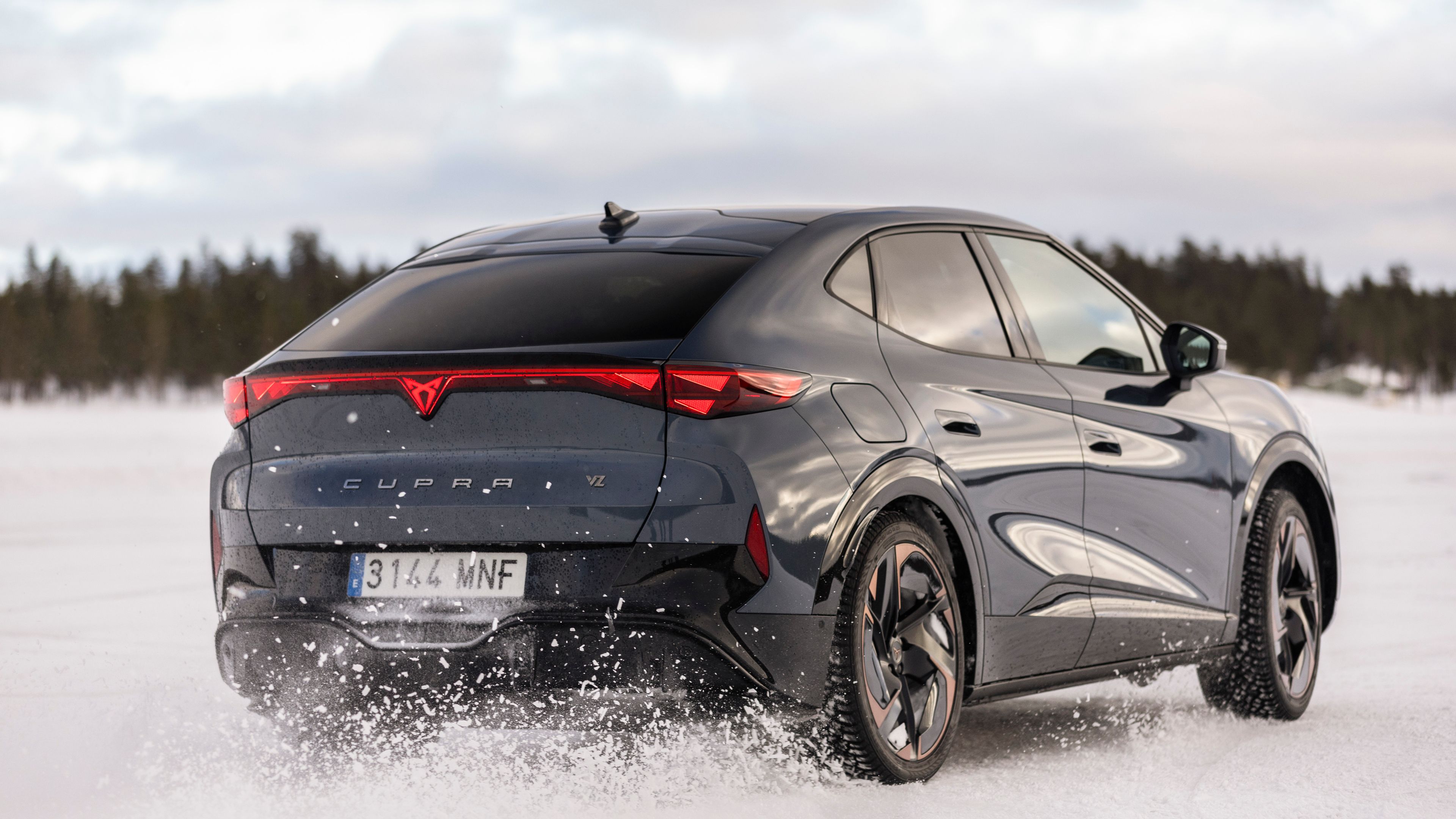 Črni športni avtomobil CUPRA Tavascan vozi po zasneženi pokrajini. Kolesa dvigajo sneg v zrak, kar ustvarja dinamičen učinek gibanja. V ozadju so vidna zasnežena drevesa pod oblačnim nebom.