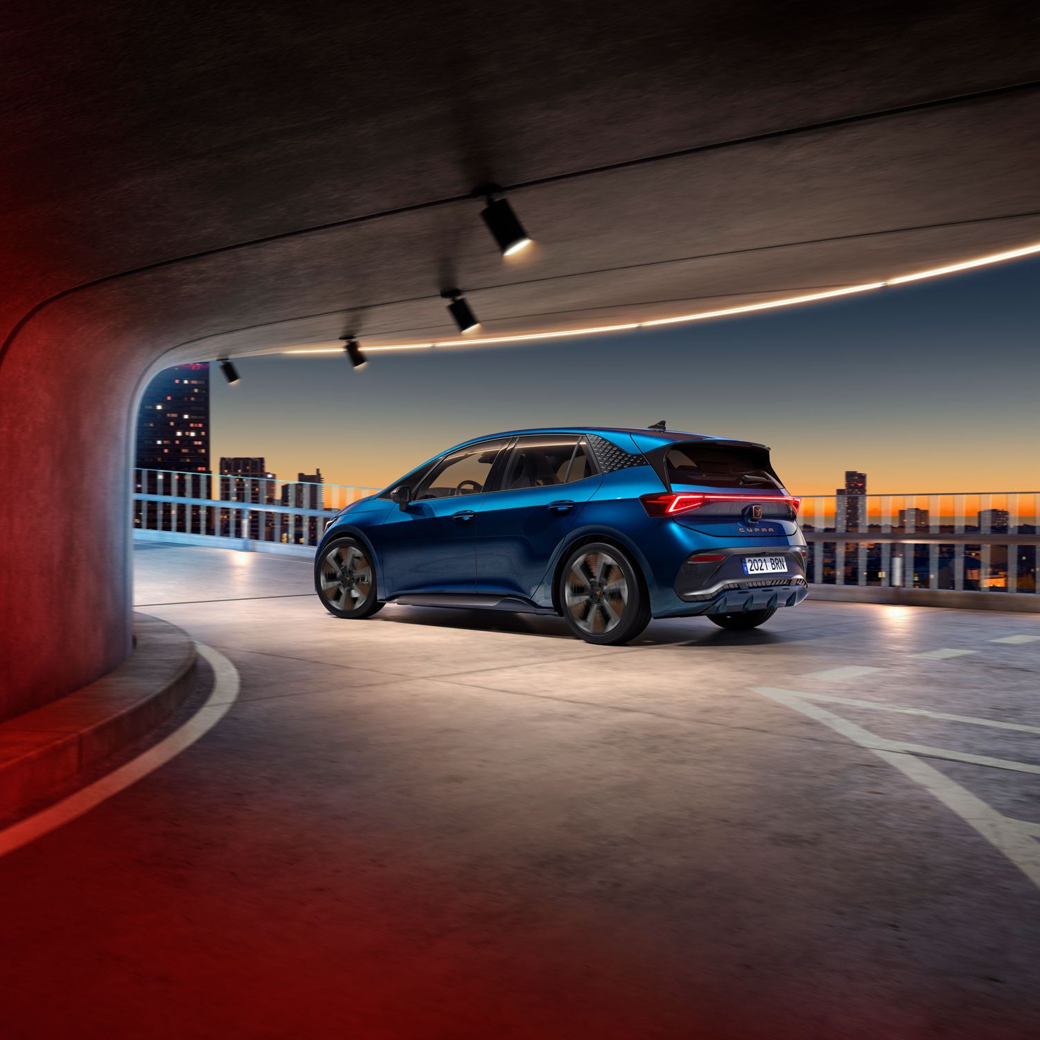 Modri CUPRA Born je parkiran v sodobni garaži z odprtim razgledom na mestno obzorje ob sončnem zahodu. Osvetlitev v garaži ustvarja toplo vzdušje.