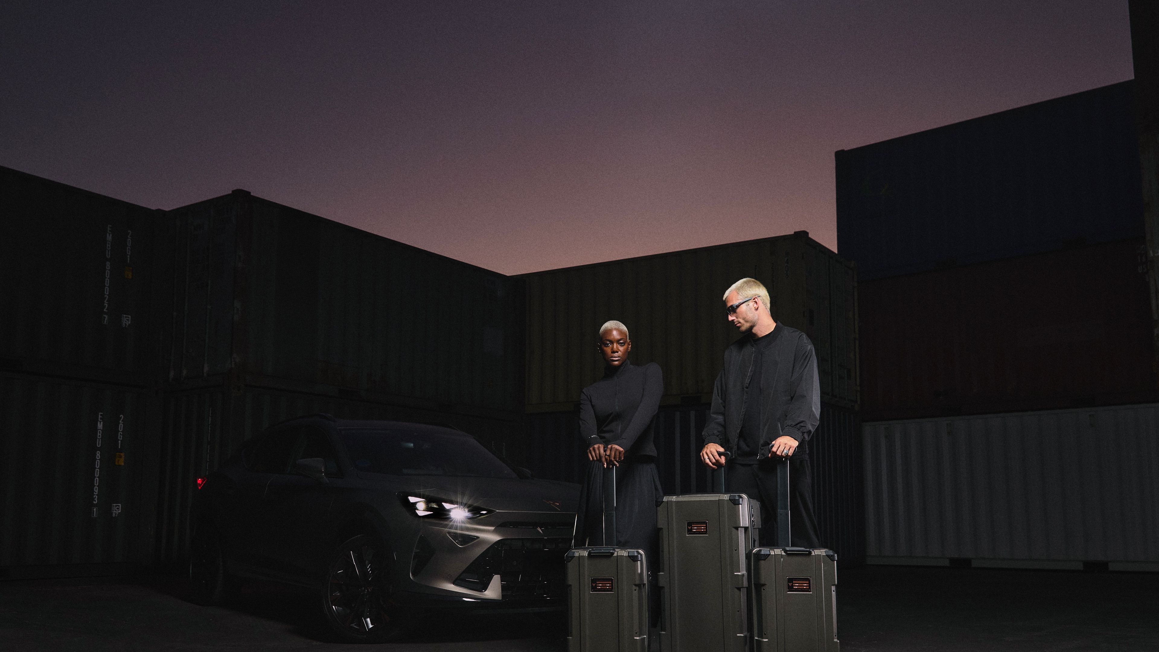 Dvije osebi stojita pred temnim avtomobilom ob pakirnih kovčkih, obkroženi s kontejnerji med sončnim zahodom.