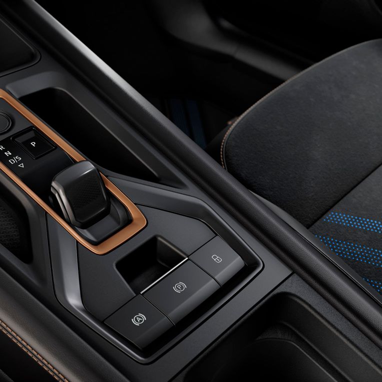 Osrednja konzola avtomobila Cupra Leon z modernimi detajli in oranžnim obrobnim videzom. Vidna so številna stikala in gumbi, vključno s prestavno ročico in elektronsko parkirno zavoro. Oblazinjenje je črne barve in vključuje modre linijske dodatke.